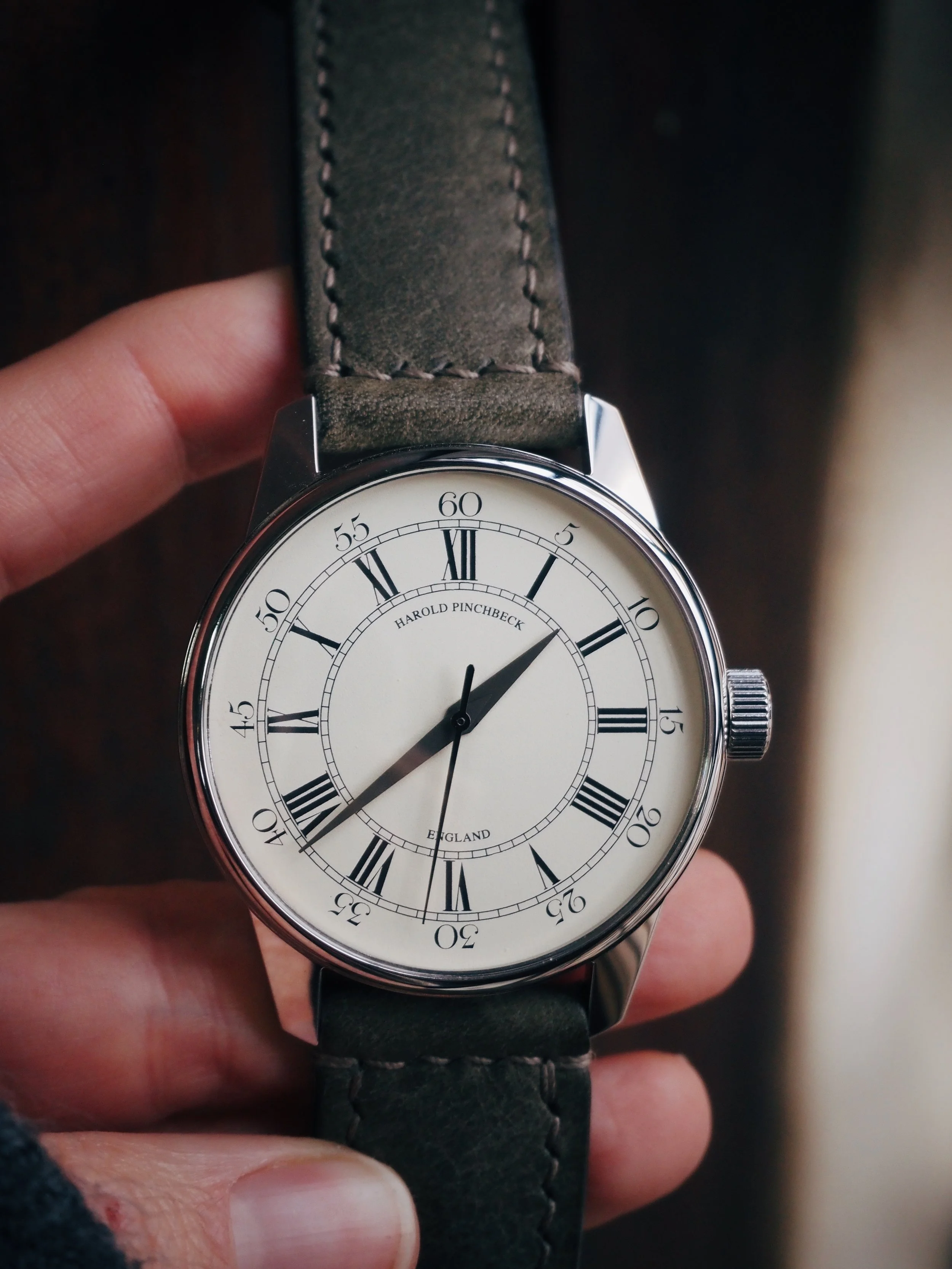 A person holding a Harold Pinbeck wristwatch with a white face, black Roman numerals, and black hour and minute hands, wearing a dark leather strap.