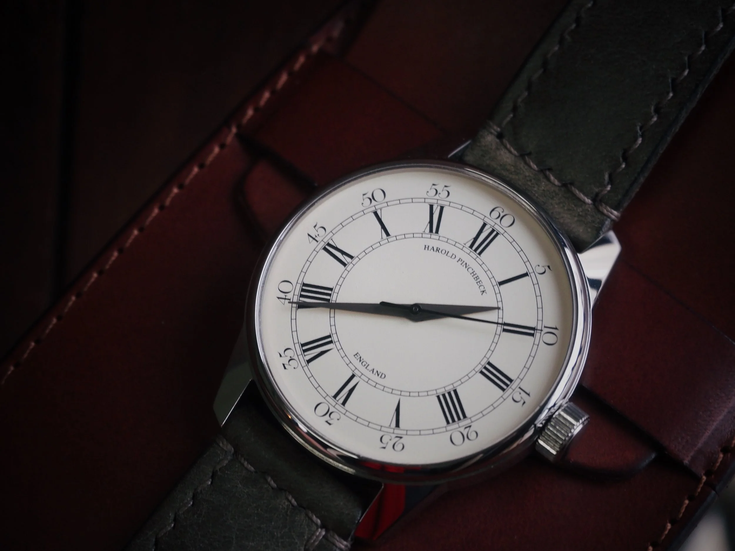 A Harold Pinchbeck English hand-made wristwatch with a white face, black Roman numeral hour markers, and black hands, showing the time as approximately 9:13, rests on a brown leather surface.