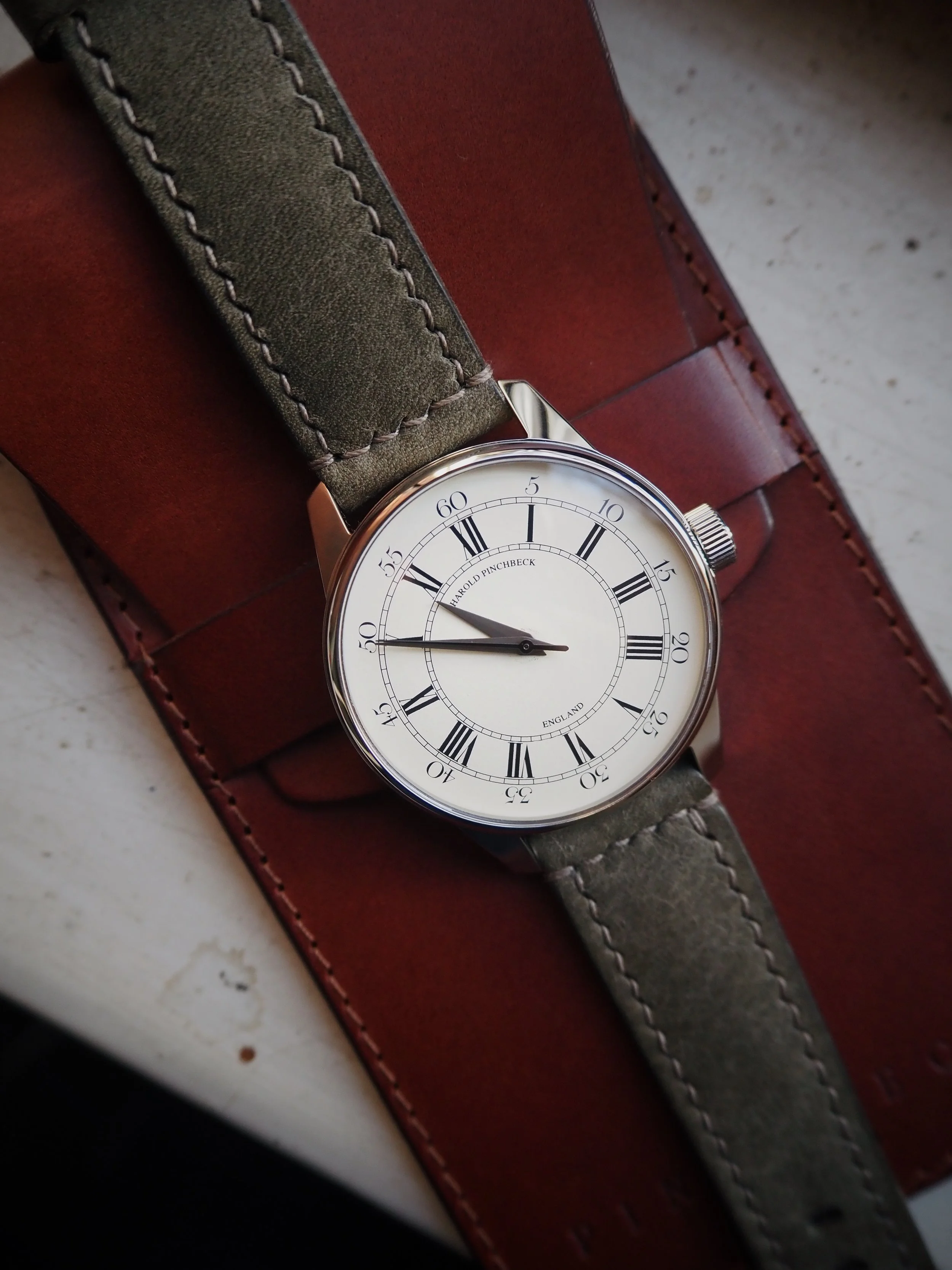 An analog wristwatch with a white dial and black Roman numerals, showing the time as approximately 10:50, resting on a leather surface with a green fabric strap.