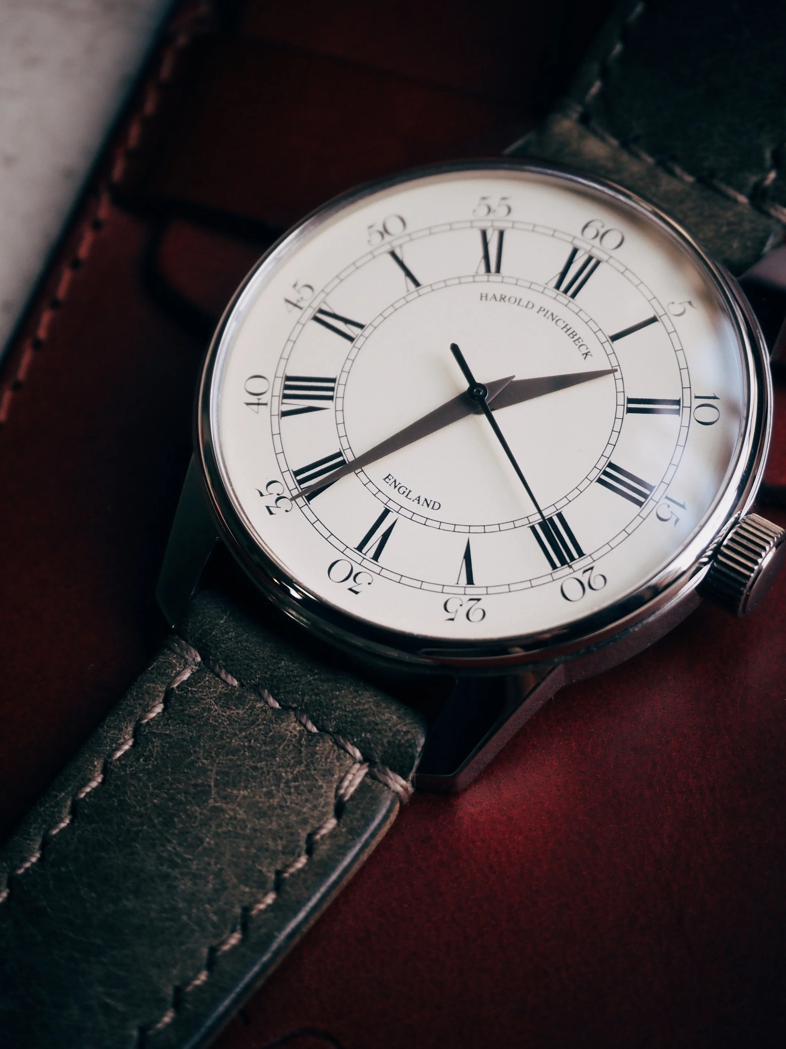 A close-up of a vintage-style wristwatch with a white face, black Roman numerals, and black minute markers. The watch has black hour and minute hands, with the minute hand pointing to 14 minutes past the hour. The strap is dark leather with visible s