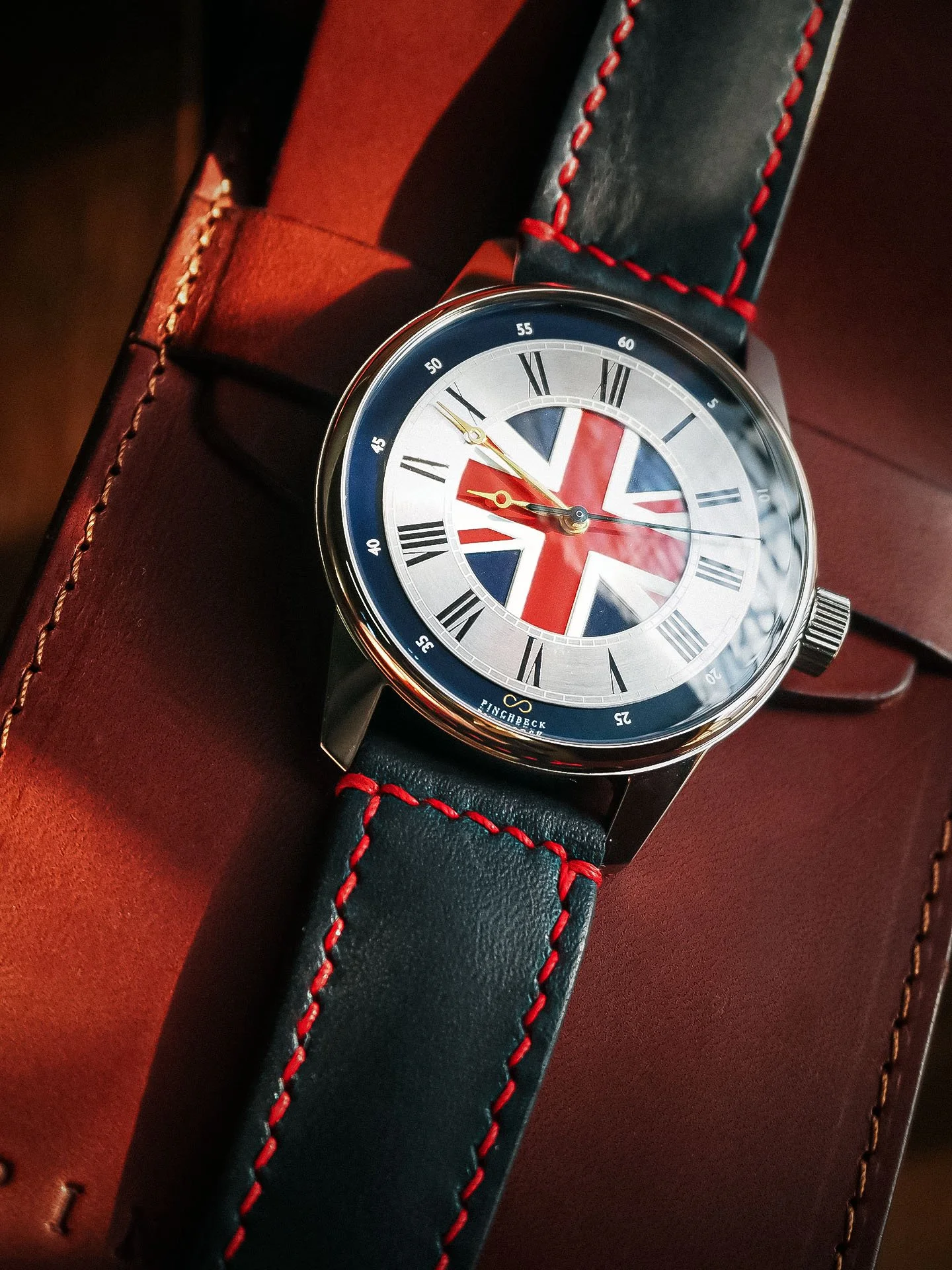 A wristwatch with a black leather strap with red stitching, featuring a face with a Union Jack design, Roman numerals, and an inset logo at the bottom reading 'Pinchbeck'.