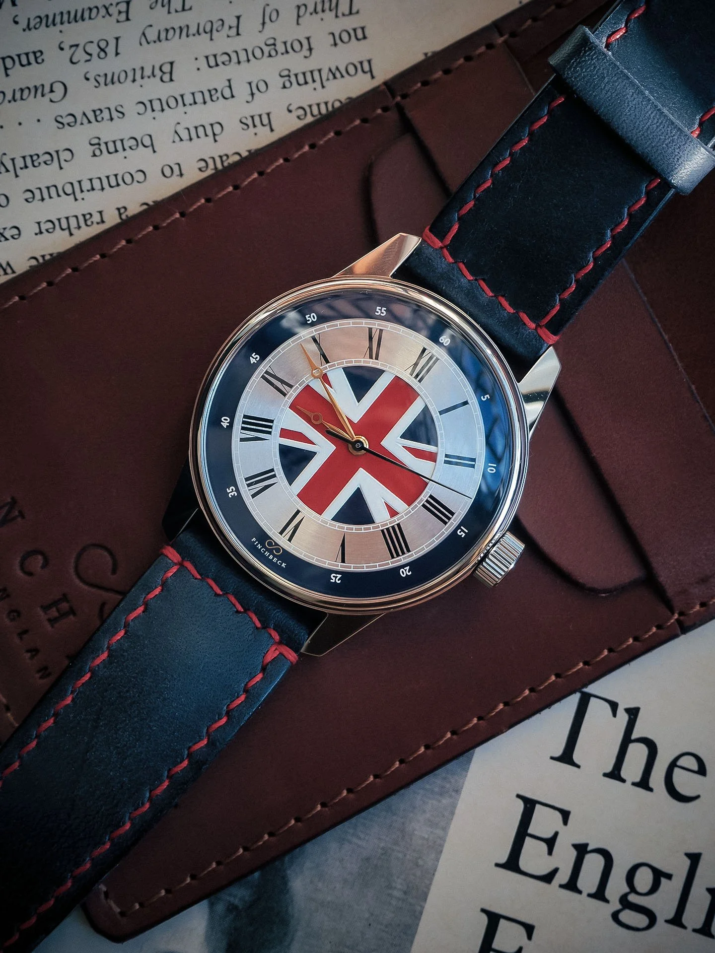 The Pinchbeck Albion watch with a dark blue leather strap with red stitching, featuring a Union Jack flag design on its dial, resting on a brown leather watch wallet and a piece of paper with printed text.