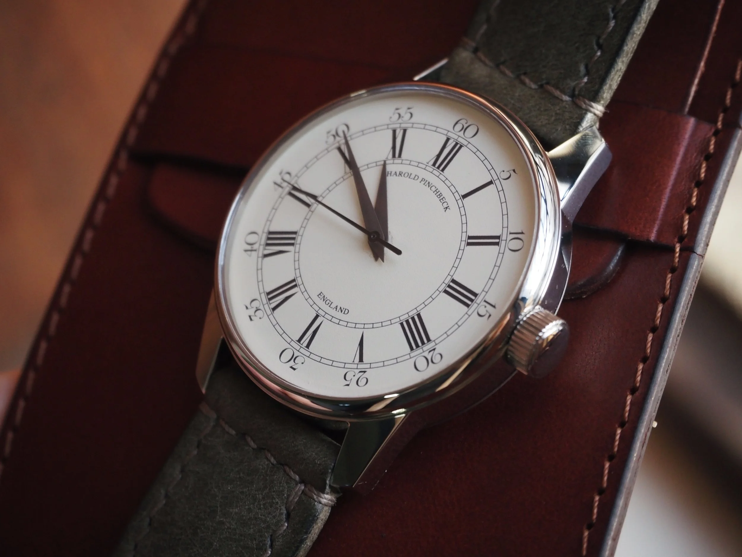 Close-up of a Harold Pinchbeck wristwatch with a white dial, Roman numerals, and a black leather strap resting on a brown leather wallet.