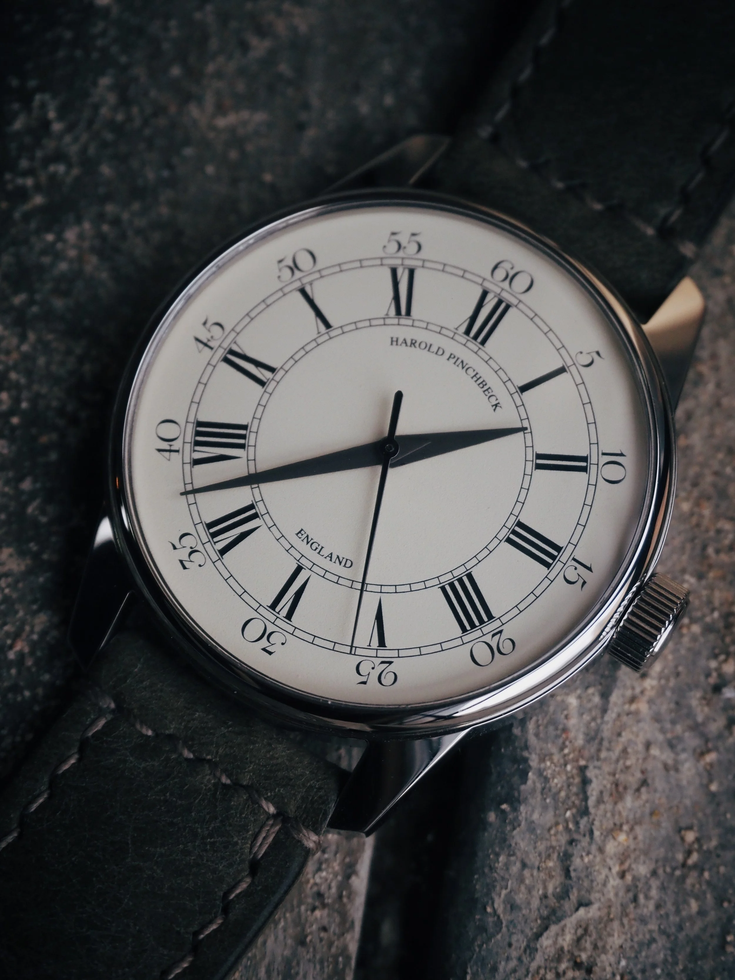 Close-up of a Harold Pinchbeck wristwatch with a white face, black Roman numerals, and a black leather strap, resting on a textured gray surface.