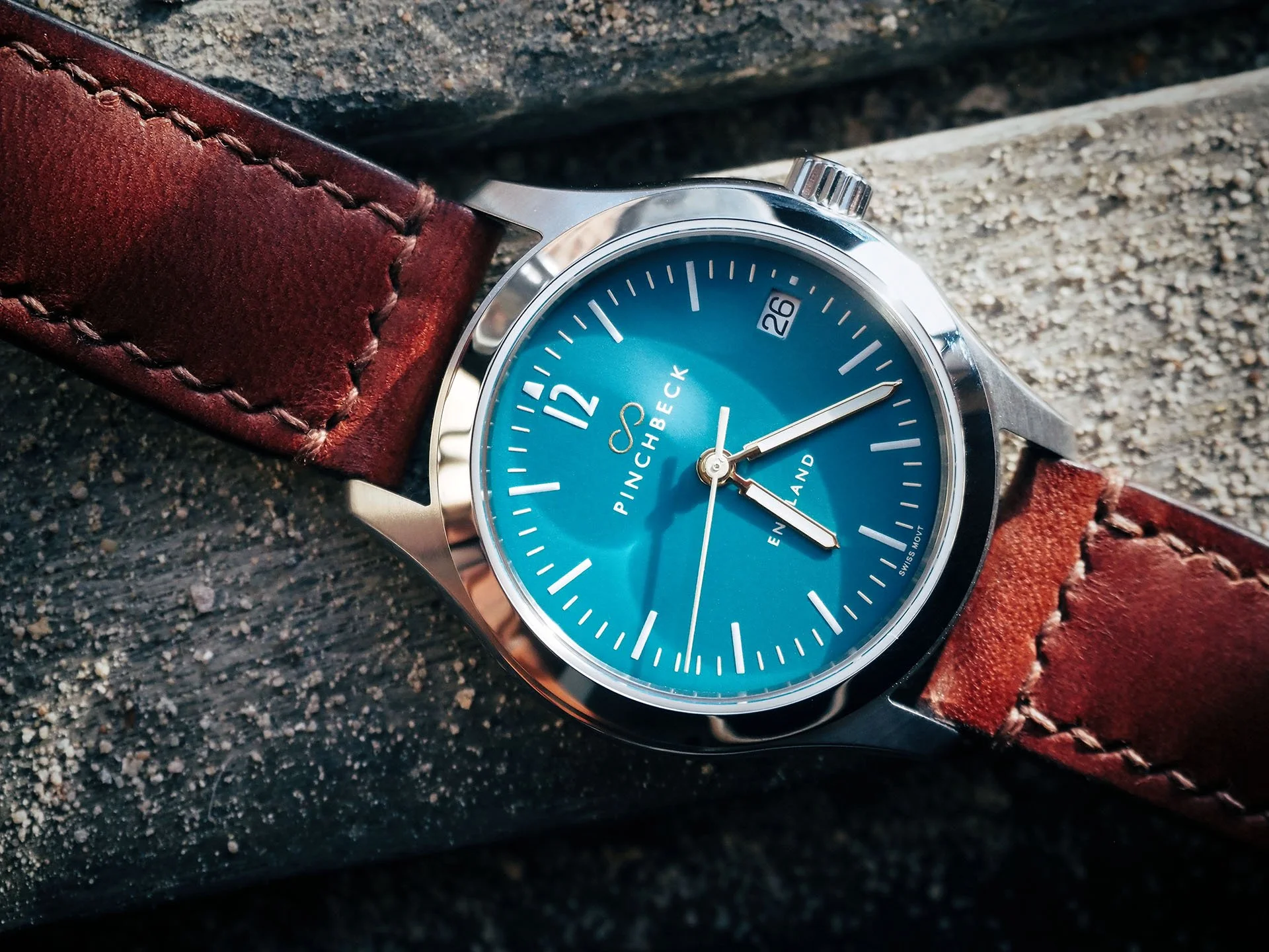 A Harold Pinchbeck Sigma wristwatch with a teal dial, stainless steel case, and hand-made Tanner Bates Conker leather strap resting on a textured surface.