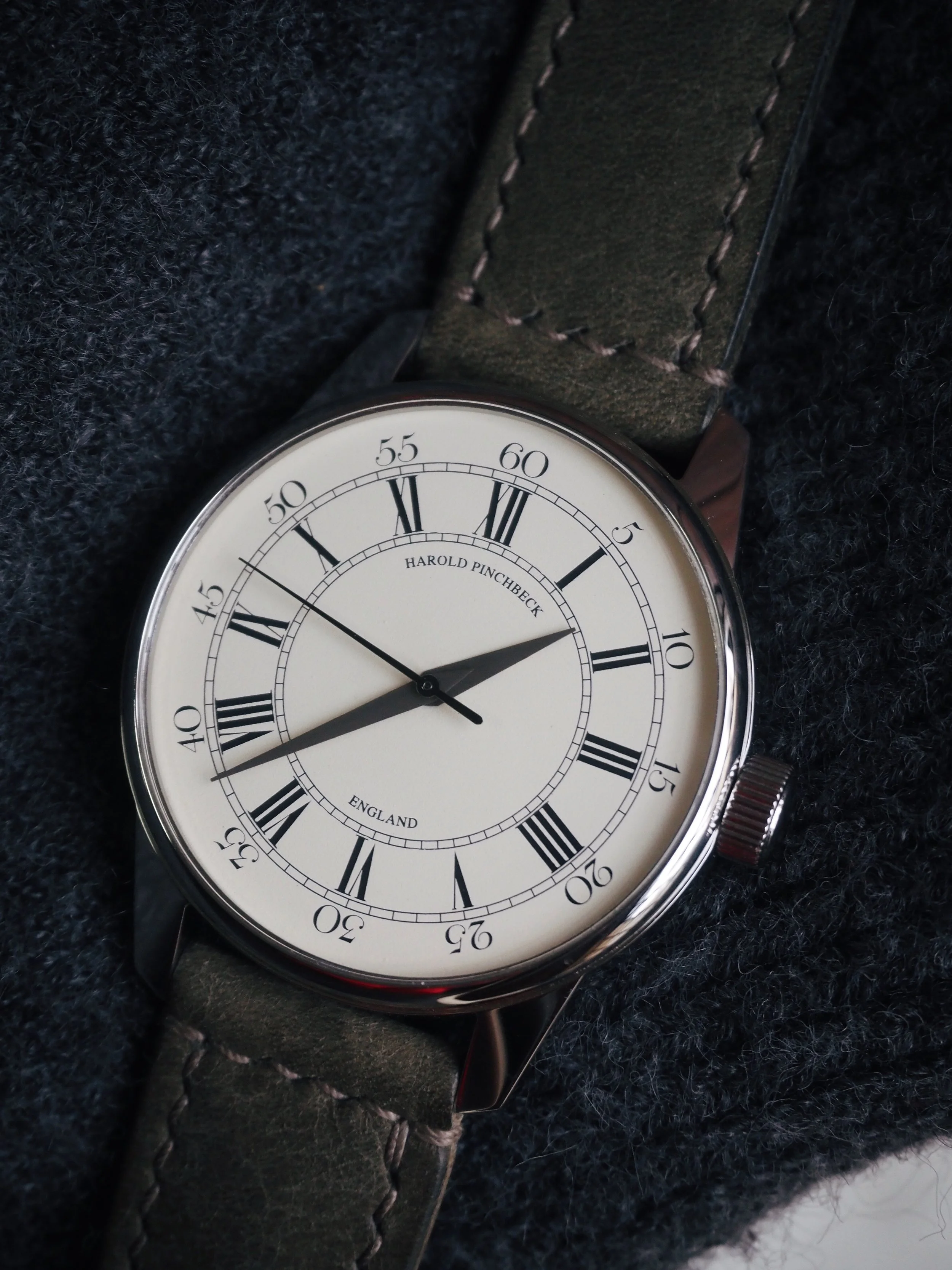 A Harold Pinchbeck wristwatch with a white dial, Roman numerals, and black hour, minute, and second hands, resting on a dark fabric surface.