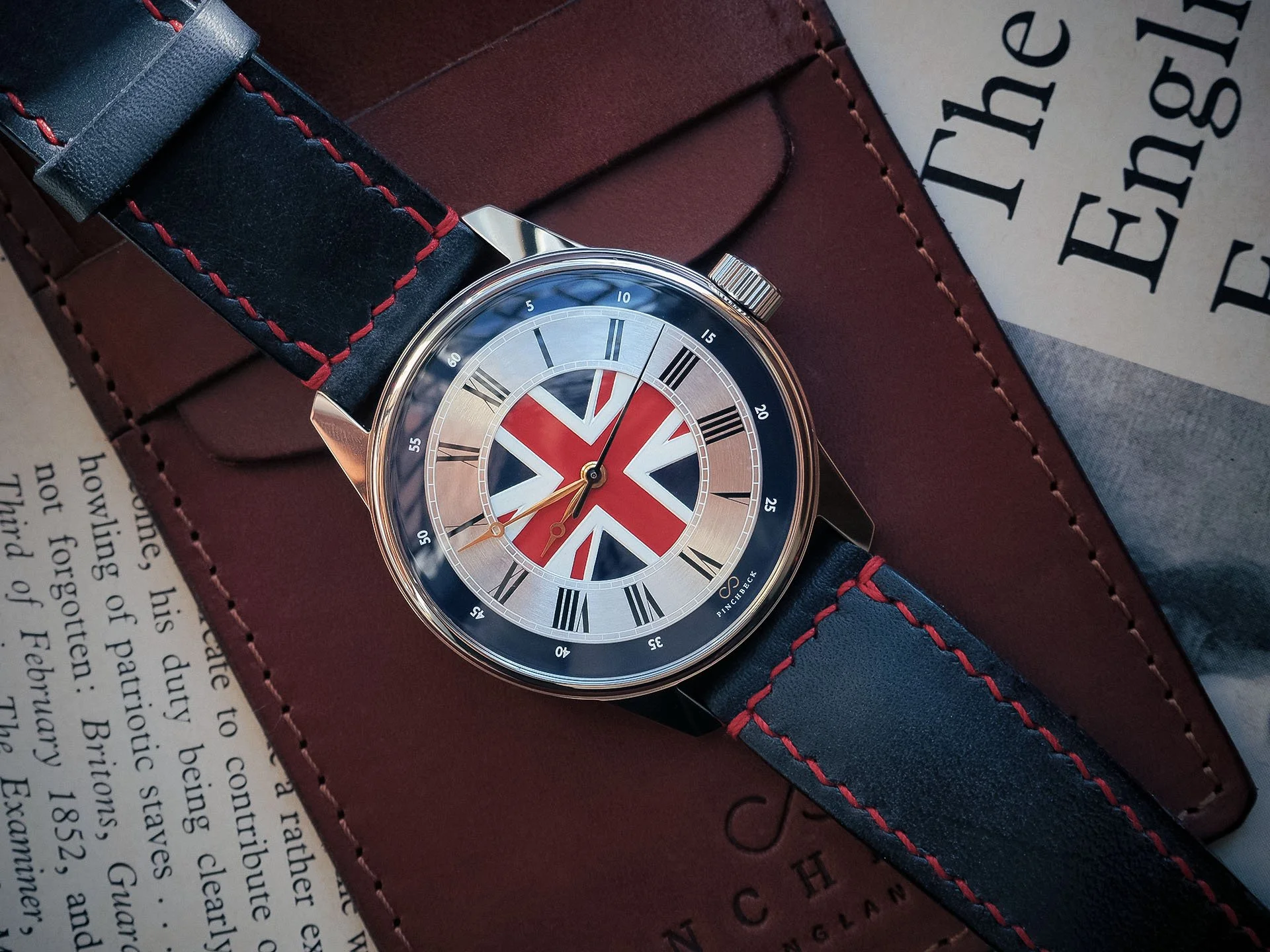 The Pinchbeck Albion watch with a dark blue leather strap with red stitching, featuring a Union Jack flag design on its dial, resting on a brown leather watch wallet and book pages with printed text.