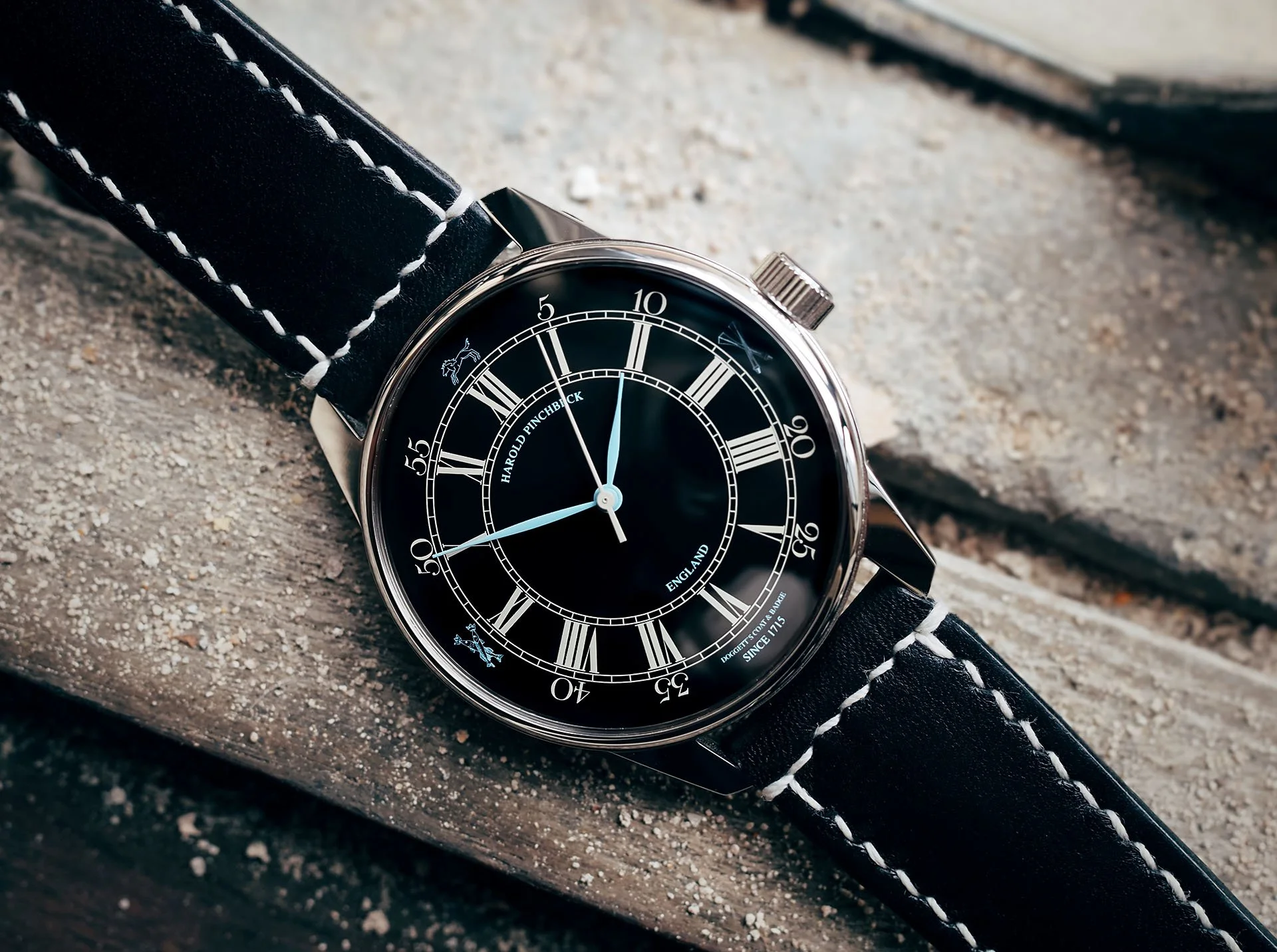 A black Harold Pinchbeck Lambeth Reach dark wristwatch with a leather strap and white stitching, displaying Roman numeral hour markers, and a black dial with blue and white accents, resting on a concrete surface.