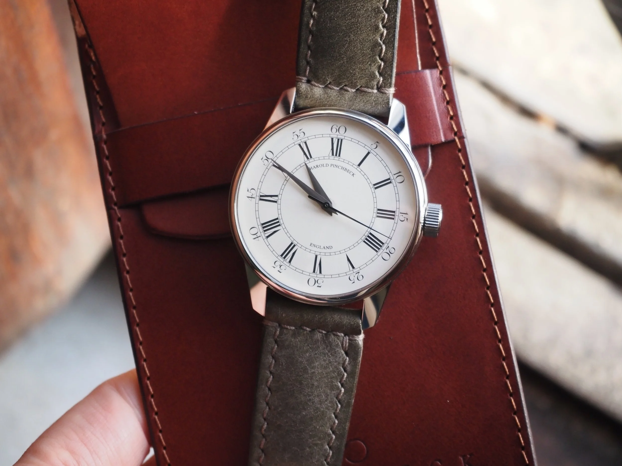 A Harold Pinchbeck watchmakers special wristwatch with a white dial, Roman numerals, and black hands, resting on a brown leather case.