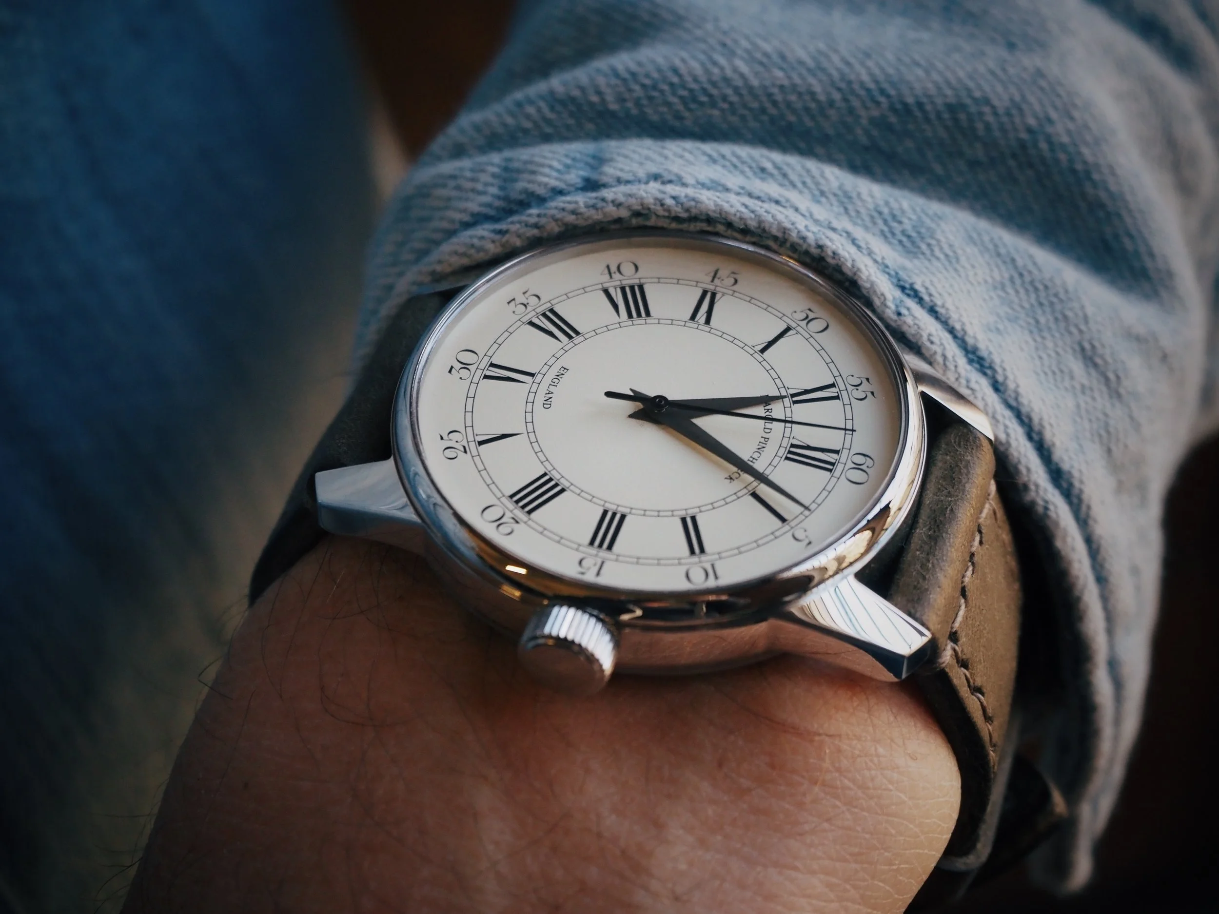 A wristwatch with a white face, black Roman numerals, and a leather strap worn on a person's wrist, with the time showing approximately 3:55.