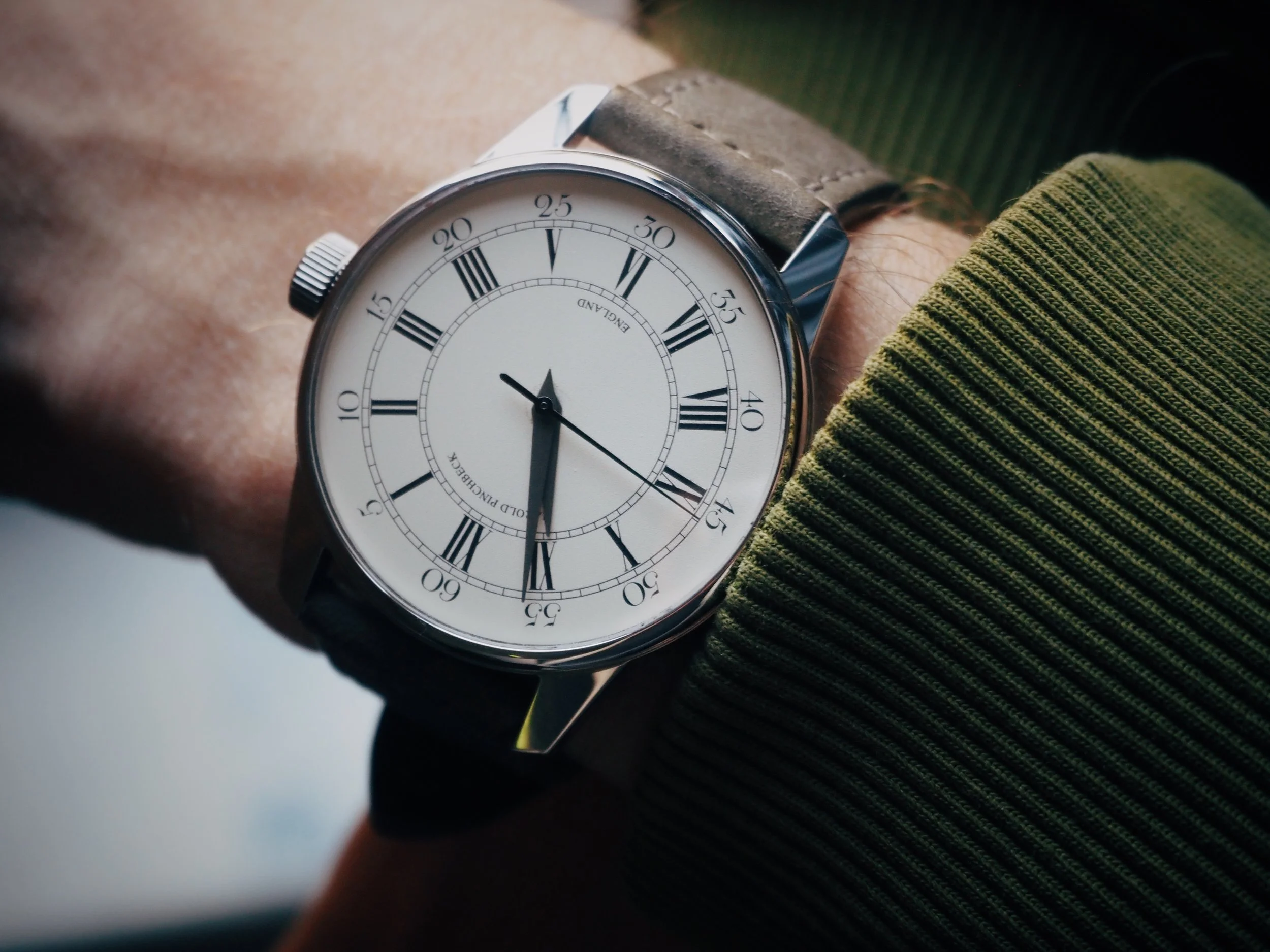 A person's wrist wearing a classic round analog watch with a white face, Roman numerals, and black hands, with a leather strap and a silver case.