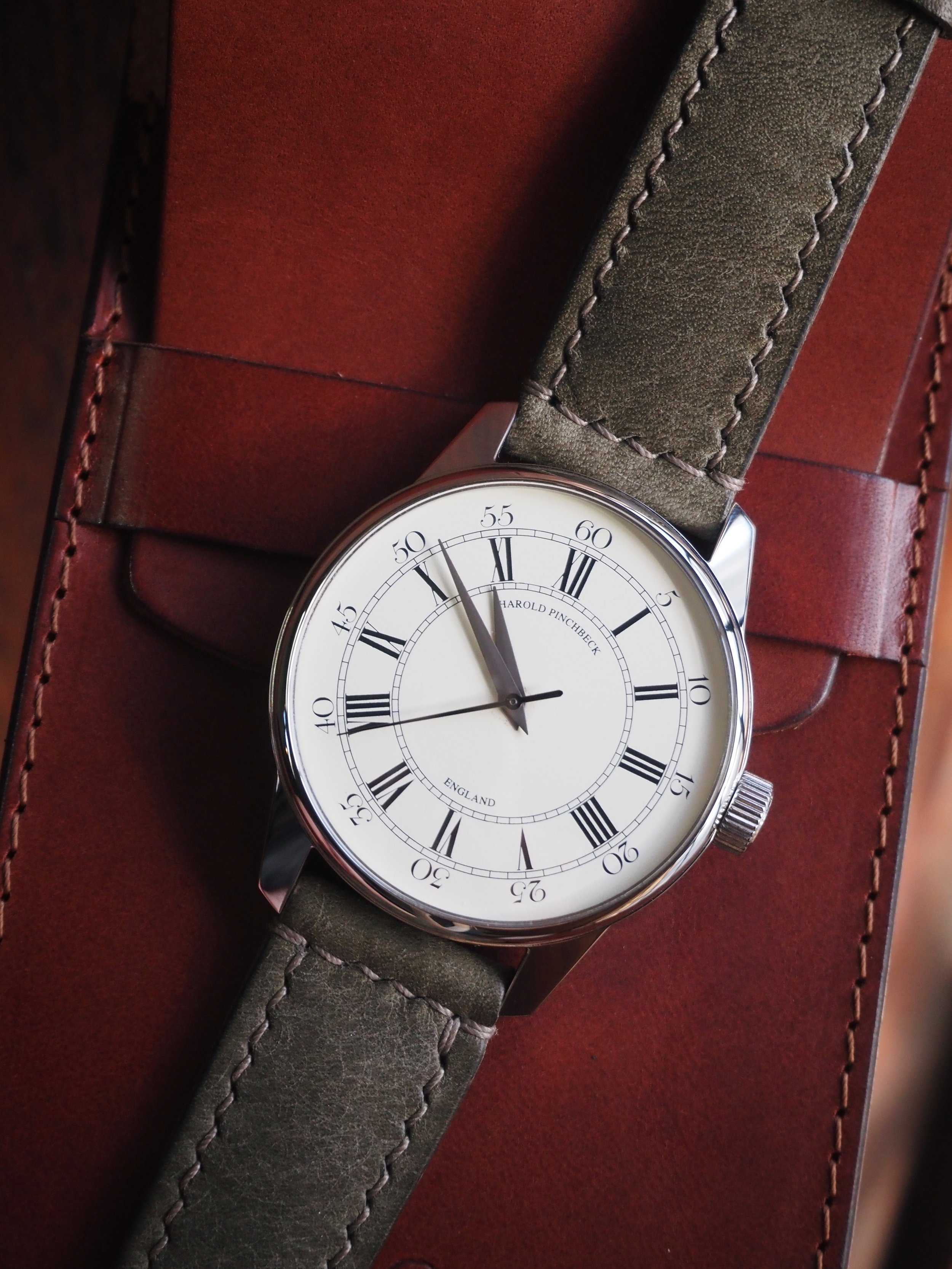 A wristwatch with a white dial, black Roman numerals, and a brown leather strap resting on a leather box.