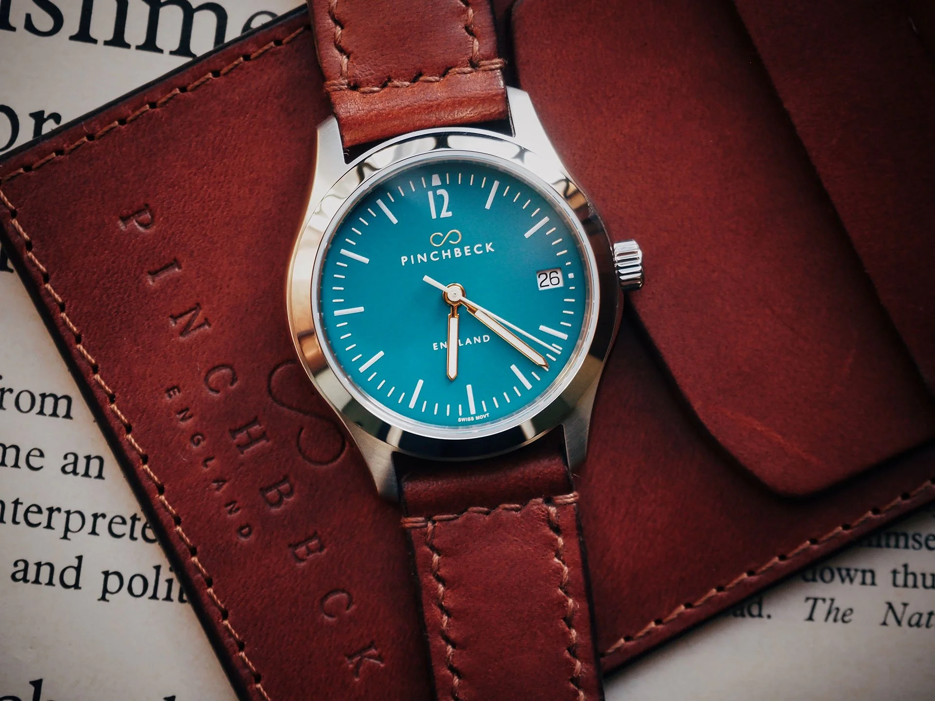 A Harold Pinchbeck Sigma wristwatch with a teal face, gold hour and minute hands, and a cream second hand resting on a red leather wallet or case. The watch displays the date as 26 and has the brand name 'Pinchbeck' and 'England' on the face.