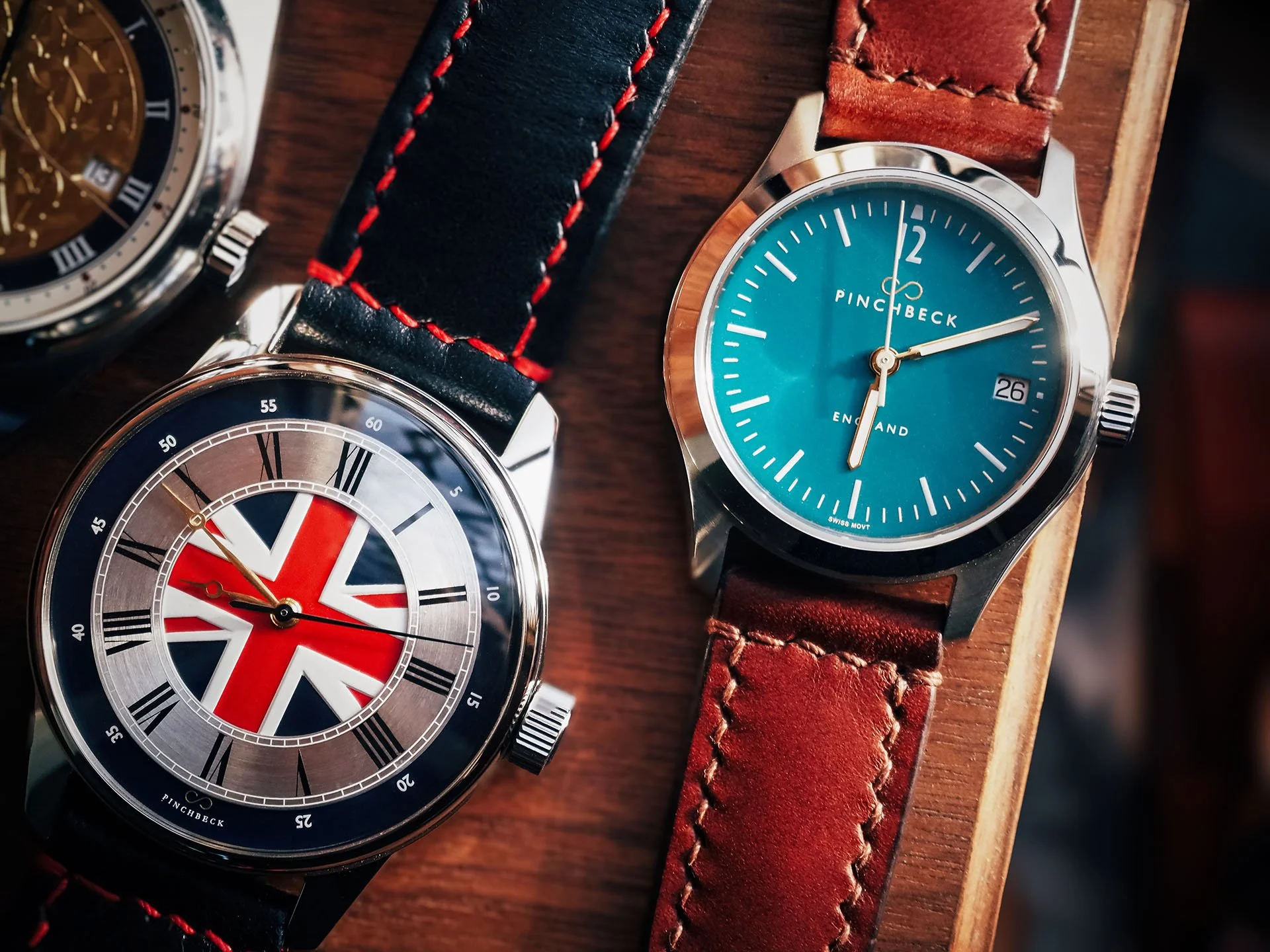 Two wristwatches on a wooden surface. The left watch features a British flag design with black Roman numeral hour markers and a black strap with red stitching. The right watch has a blue face, silver bezel, and a brown leather strap, with the time sh
