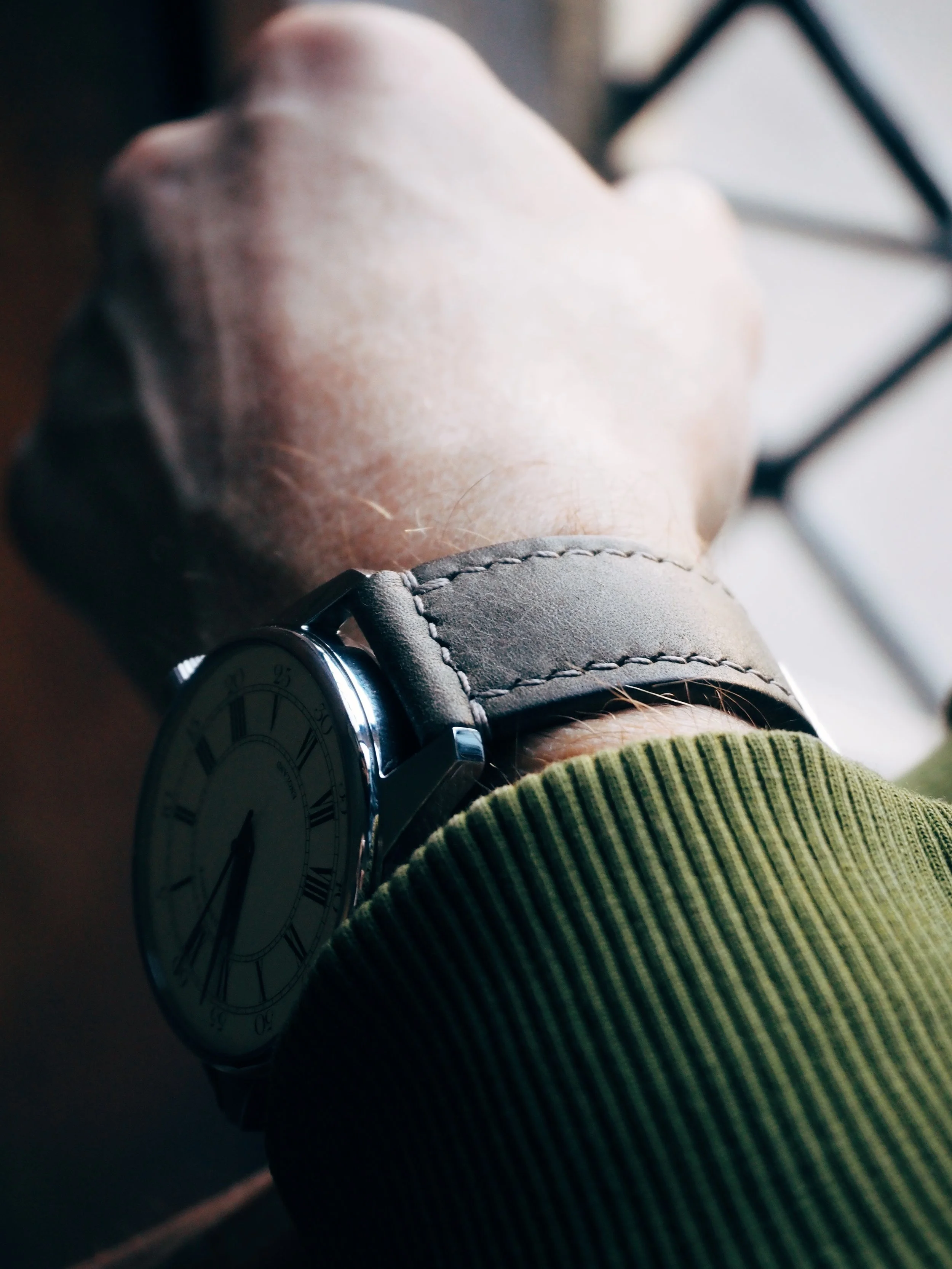Close-up of a person wearing a watch with a leather strap, seen from the wrist to the upper arm, with a green sleeve and a dark background.