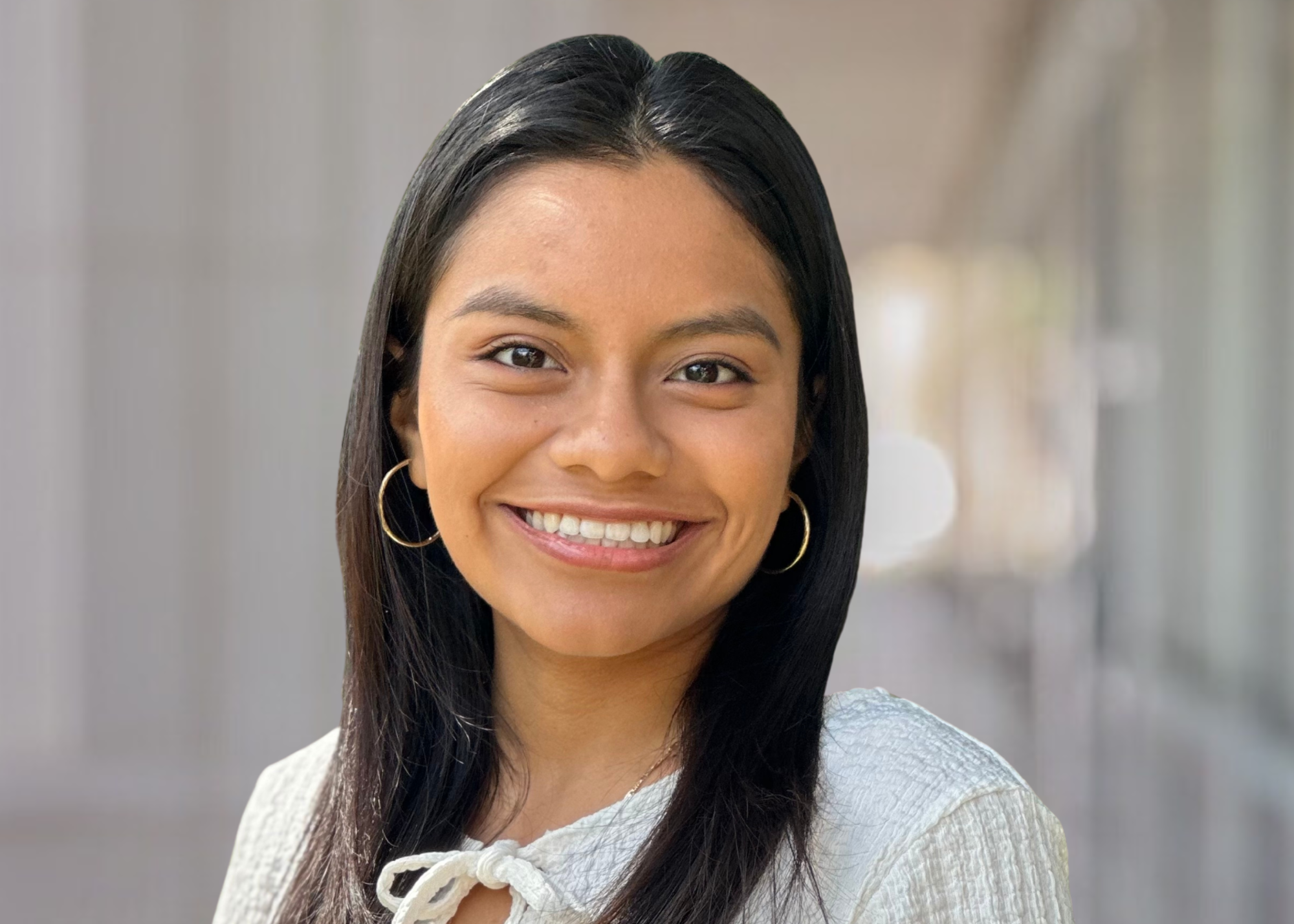 Close-up of a young woman with long dark hair, wearing a white shirt, smiling indoors.