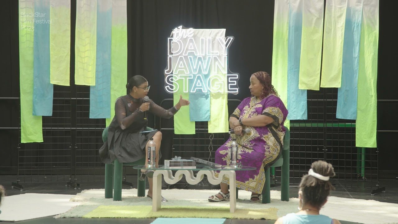 Zeinabu Davis on the Daily Jawn Stage being interviewed by Shanti Mayers. Zeinabu is wearing a purple and gold kaftan maxi dress. Shanti is wearing a black sheer midi swing dress.