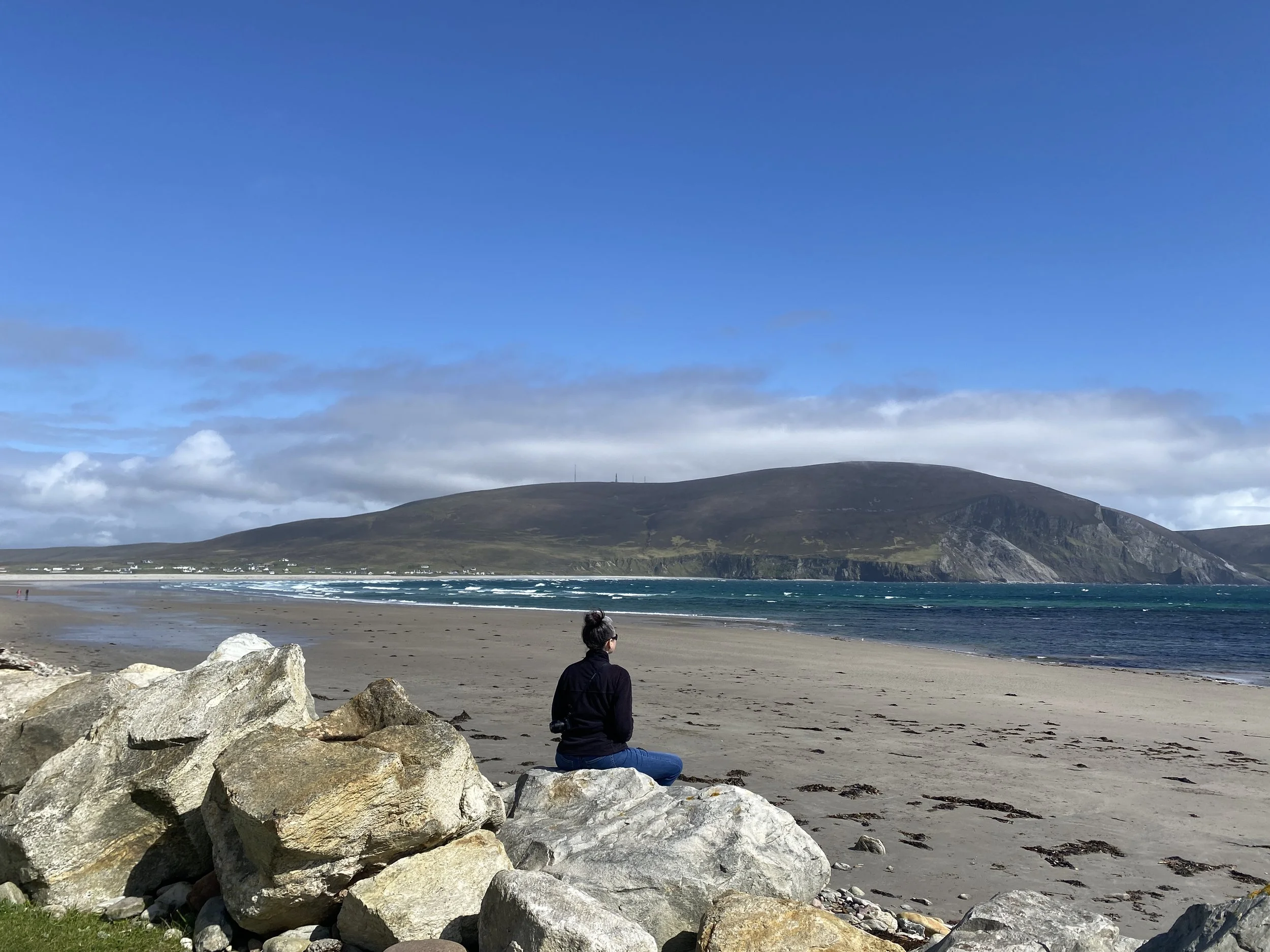 A beach scene with big rocks at the edge of the shore, looking out over the beach and mountains. A woman dressed in a black top and jeans sits on the rocks meditating peacefully.