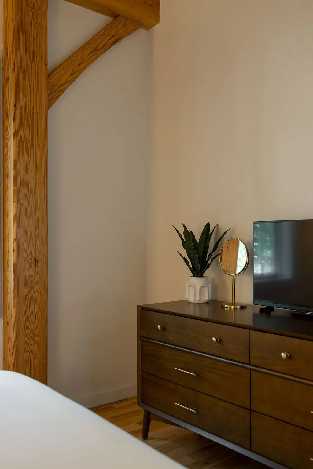 A wooden dresser with a potted plant, a small mirror, and a TV on top, in a cozy bedroom with beige walls and hardwood flooring.