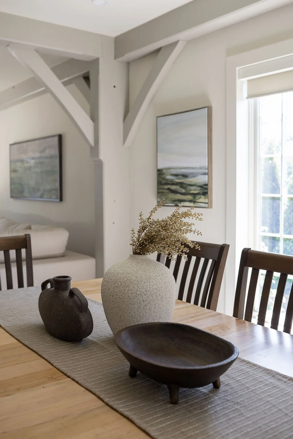 A dining room with a wooden table and a vase with flowers. On the table, there is an art sculpture and a dark wooden bowl. The room has clean fresh white walls with two landscape paintings, and a window with white blinds letting in natural light.