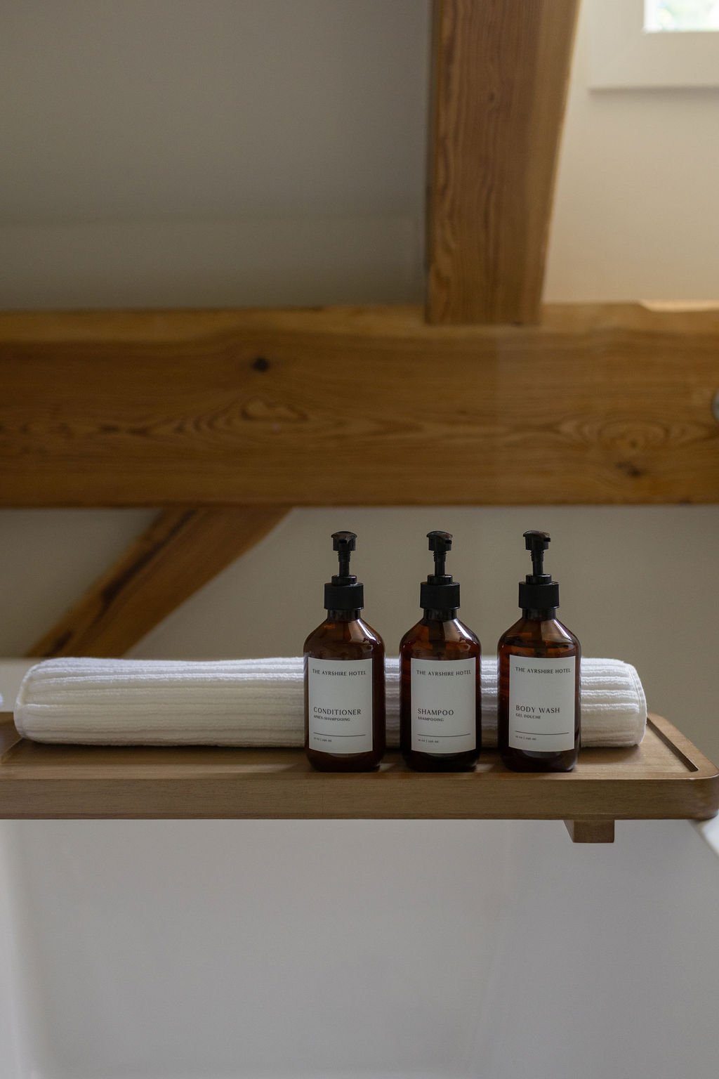 Three amber bottles filled with premium shampoo, conditioner, and body wash sit on a wooden tray, with a roll of white towels behind them.