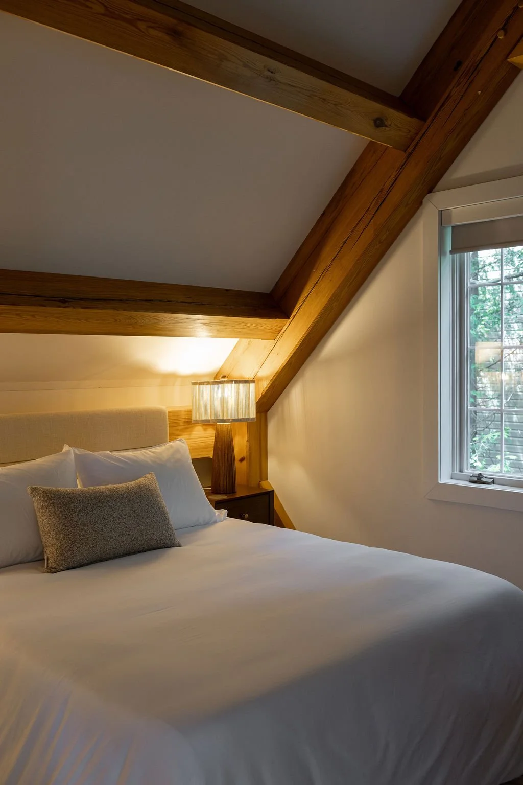Cozy bedroom with sloped ceiling, wooden beams, a bed with white bedding and pillows, a bedside lamp, and a window with view of trees outside.