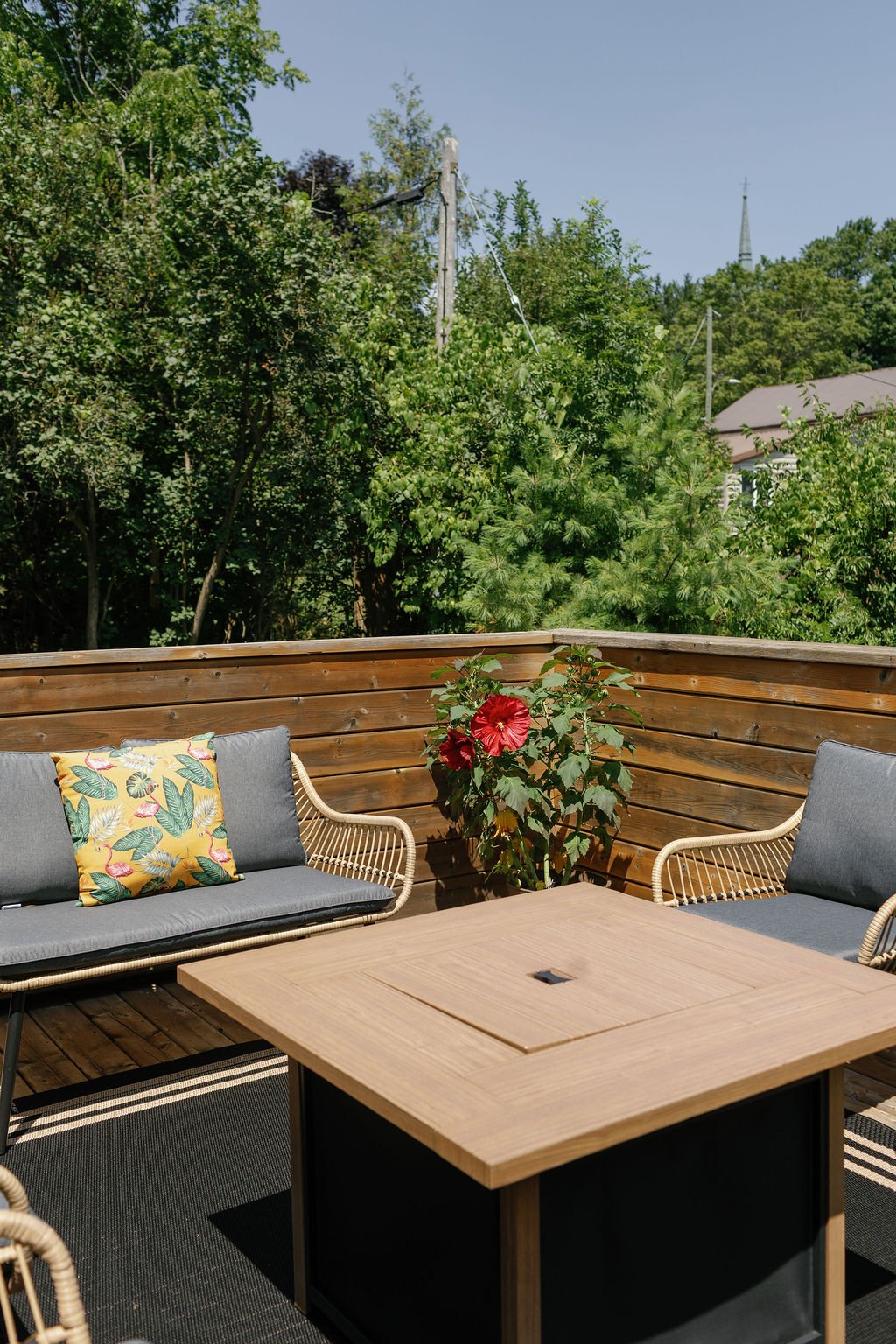 A private outdoor patio with cushioned chairs, a fire table, and a vibrant red hibiscus plant, surrounded by lush green trees and a wooden fence.