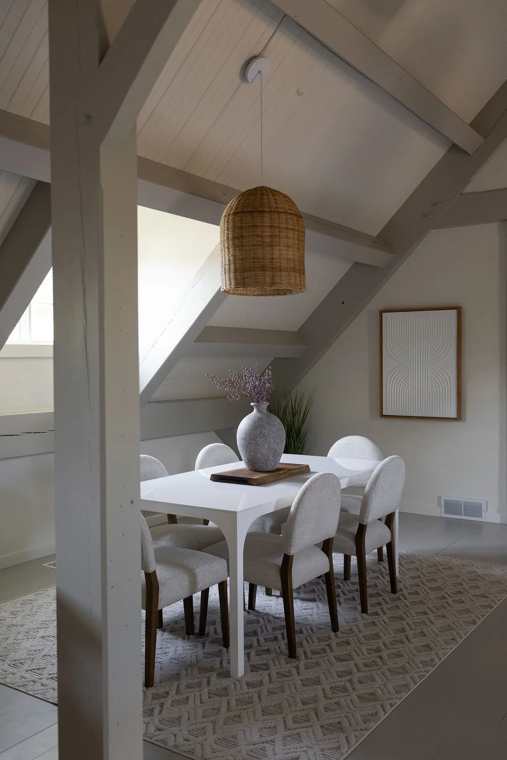 Dining room with a white table, six beige upholstered chairs, a wooden tray with a gray vase containing purple flowers, a geometric wall art piece, a wicker pendant light, a skylight, and a patterned rug.