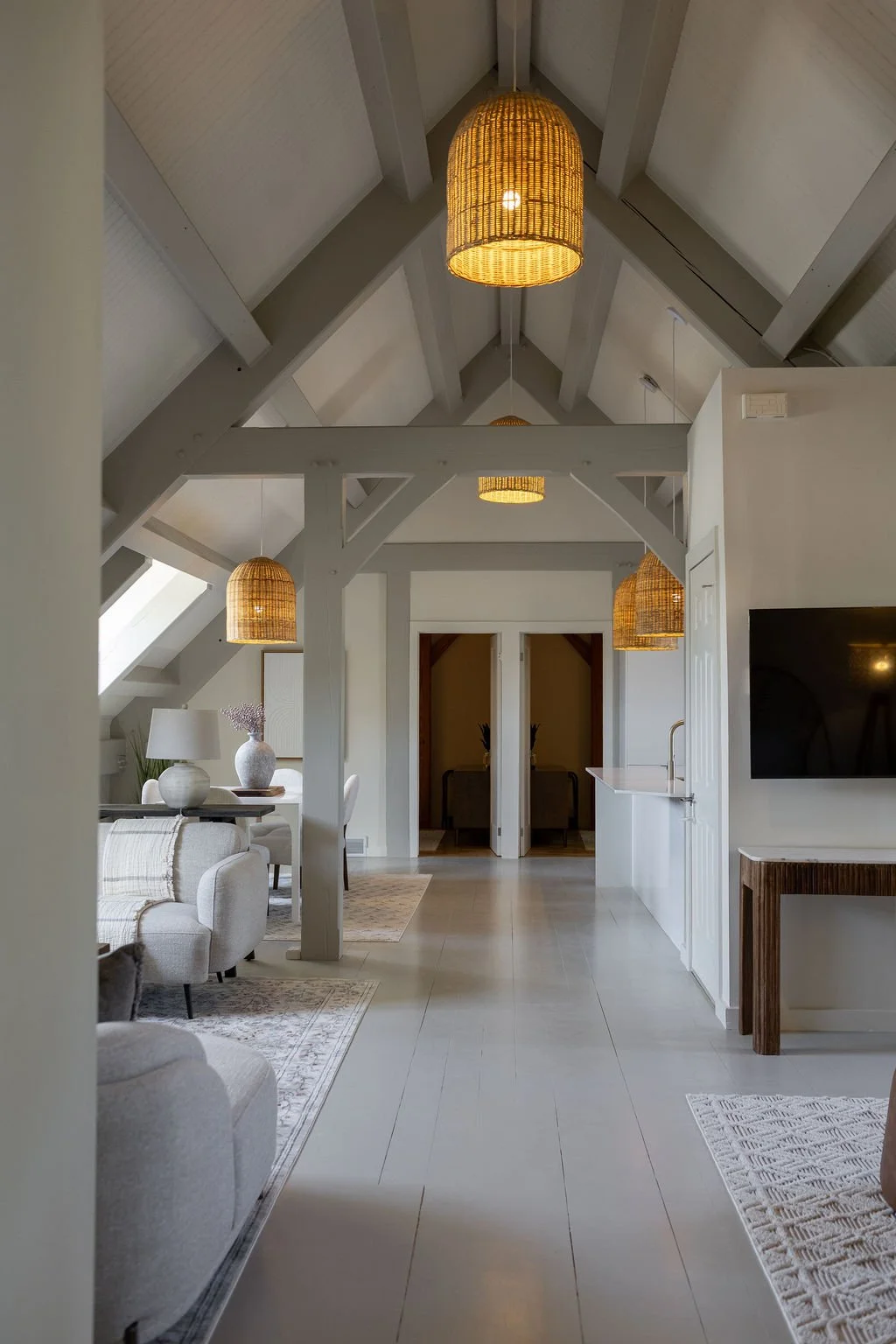 Bright second floor living room in the villa. With white walls, exposed beams, wicker pendant lights, cozy sofa, and a dining area in the background.
