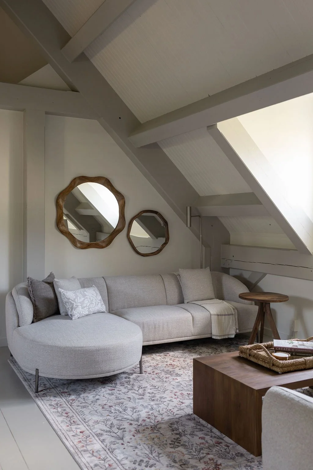Living room with a beige sectional sofa, decorative pillows, a patterned area rug, a wooden coffee table, and two wall mirrors with wooden frames under a sloped ceiling.