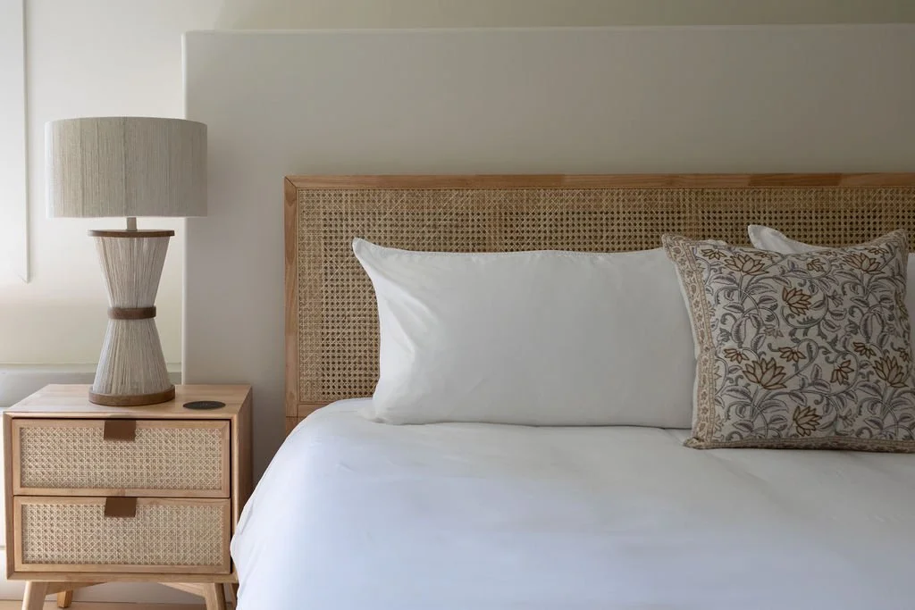 A neatly made king sized bed with white pillows and a decorative pillow, a wooden headboard, and a wooden nightstand with a lamp, in a modern hotel bedroom with light-colored walls.