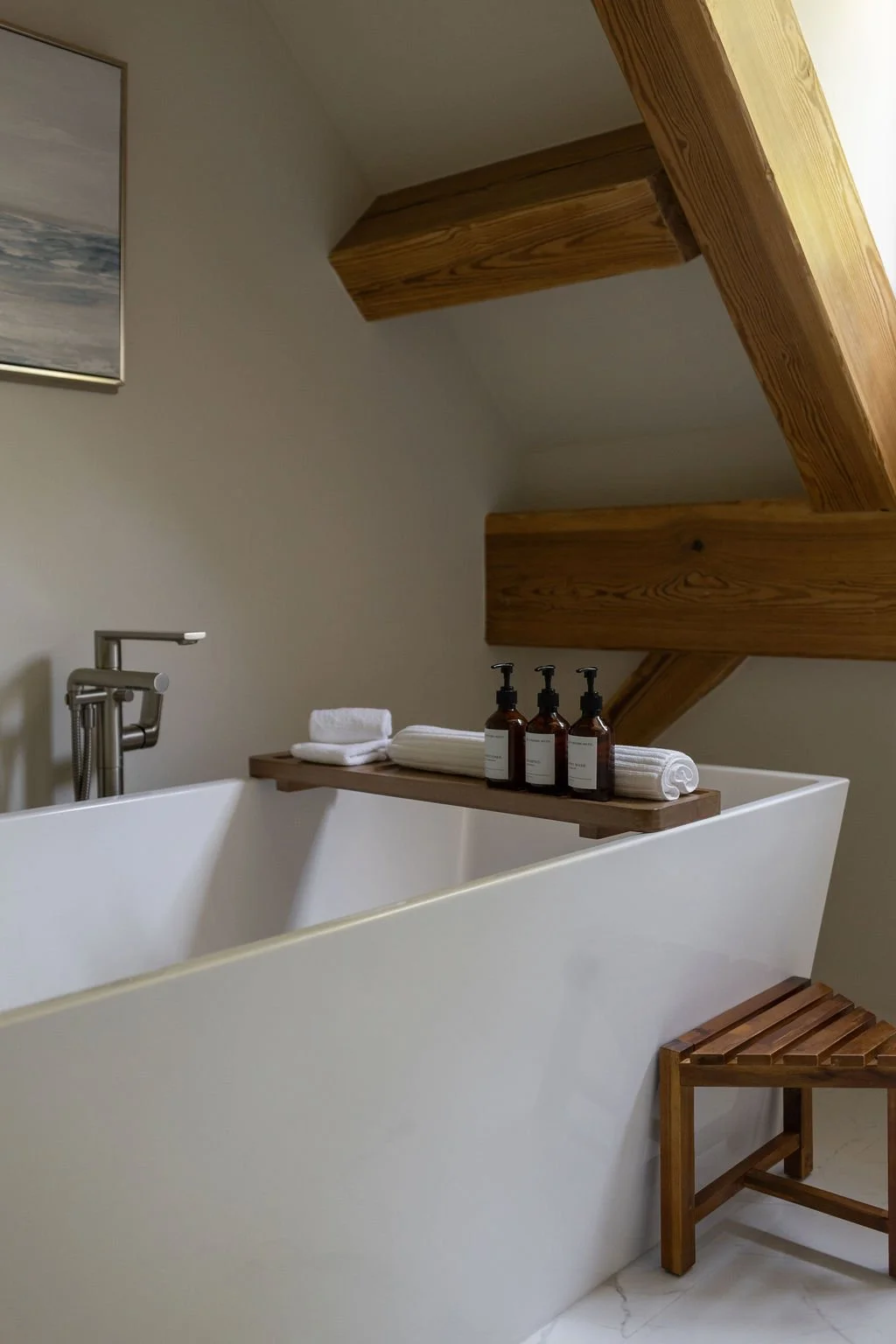 A modern, minimalistic bathroom with a white soaker bathtub, a wooden shelf holding towels and toiletries, and exposed wooden ceiling beams.
