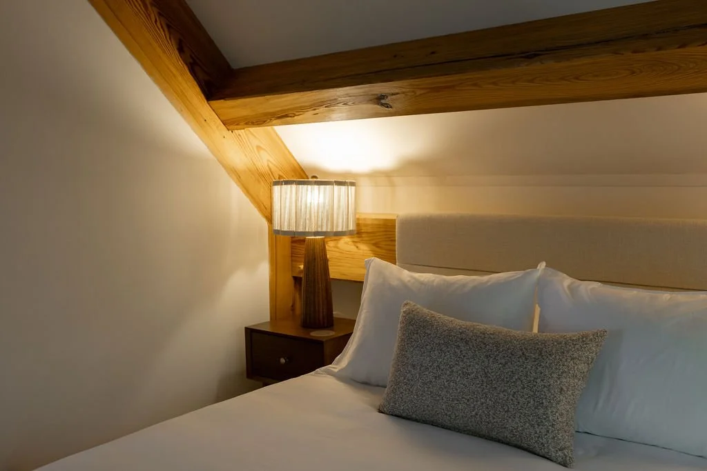 A bedroom with a bed, white pillows, a table lamp, and wood beams in the ceiling.