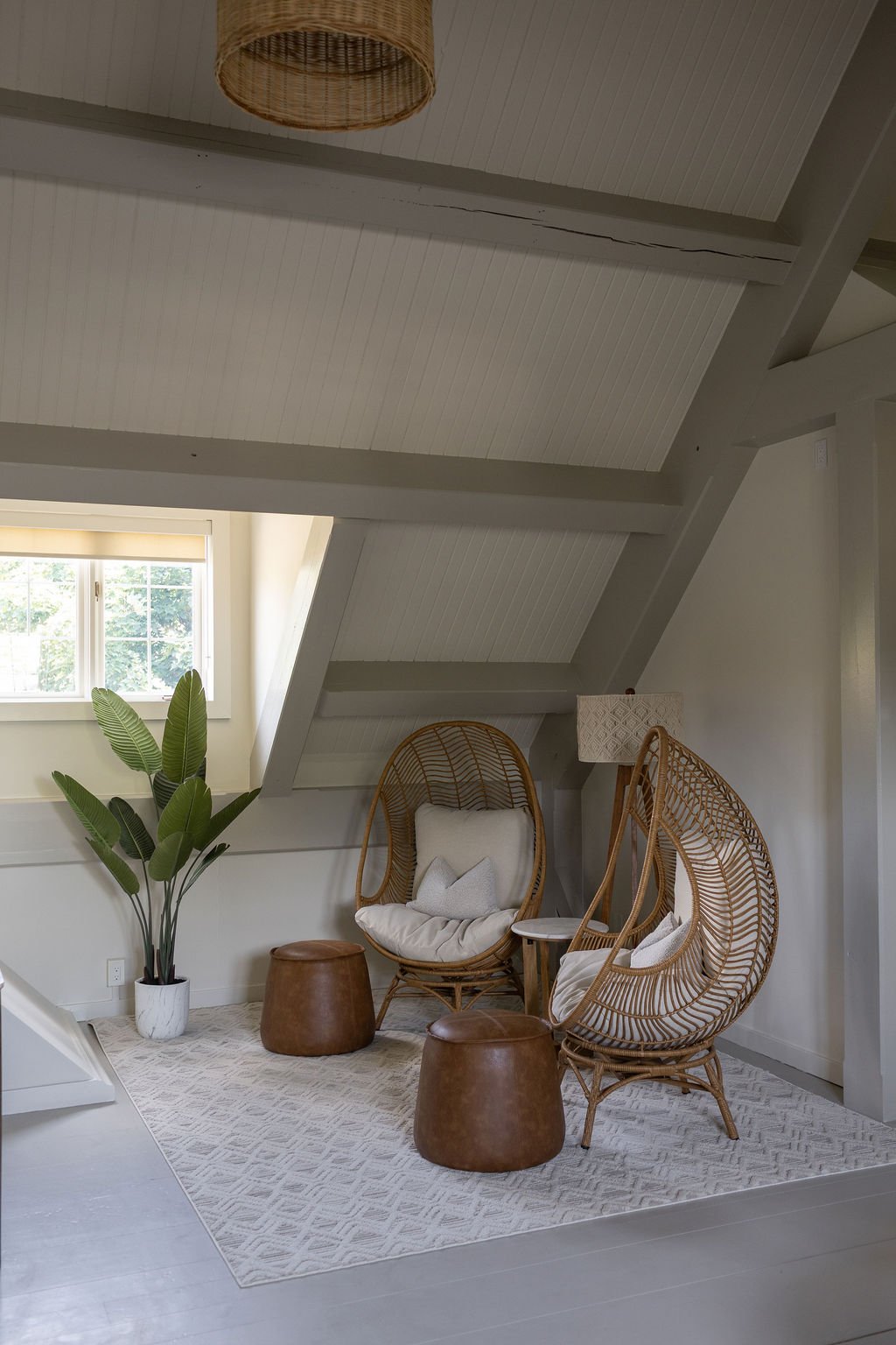Cozy seating area with two rattan egg chairs, white cushions, a small round side table between them, two leather ottomans, a woven floor rug, and a window with natural light in under the vaulted ceilings