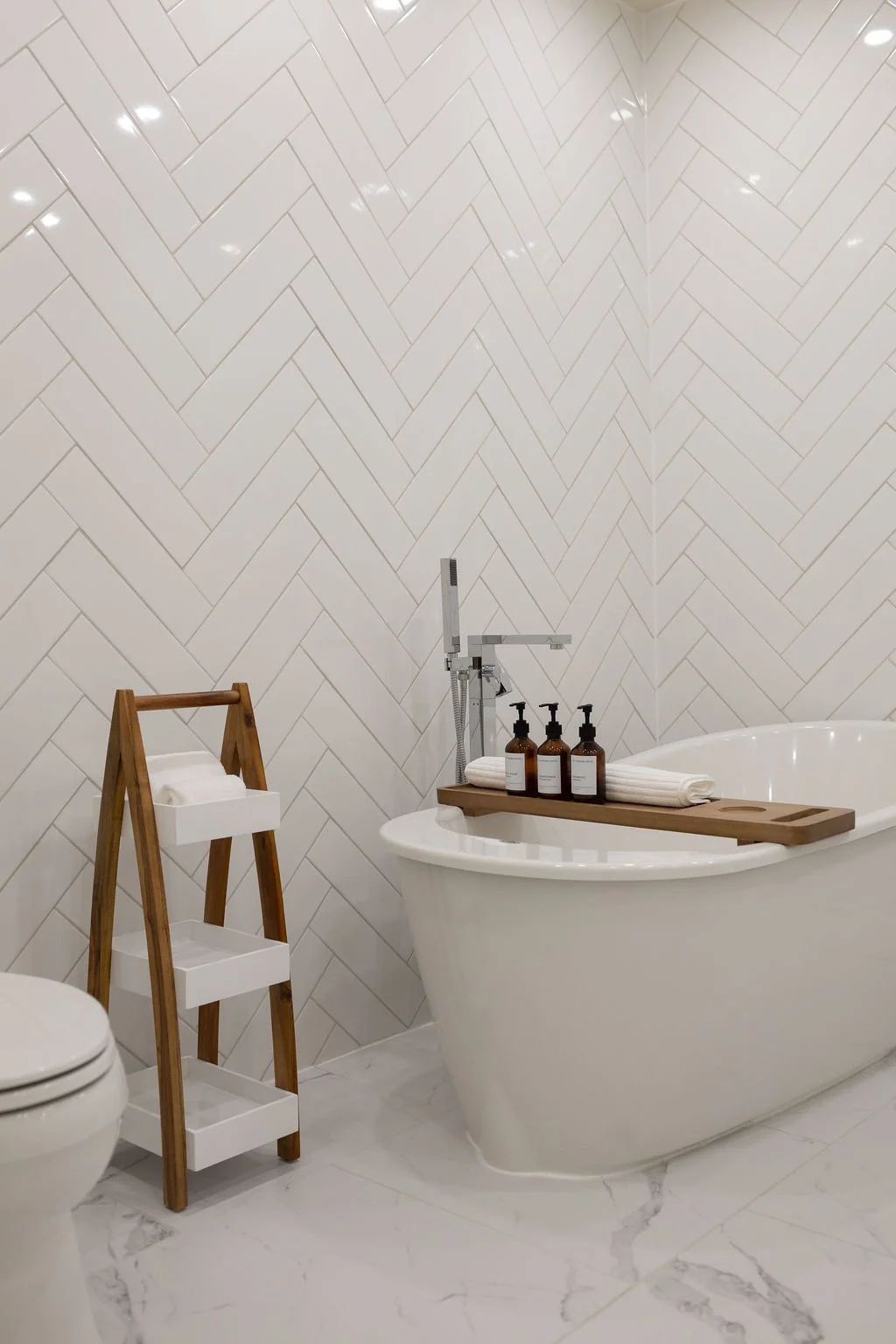A modern luxury bathroom with white herringbone-tile wall, white soaker style bathtub with wooden tray holding three premium soap bottles and rolled towels, all on a white marble floor.