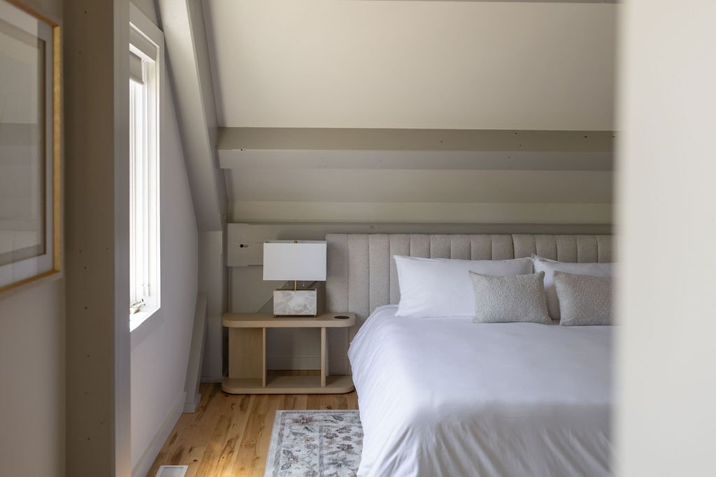 A cozy bedroom with a sloped ceiling, a beige upholstered bed with white bedding and pillows, a wooden nightstand with a lamp, a window with blinds, and a rug on a wooden floor.