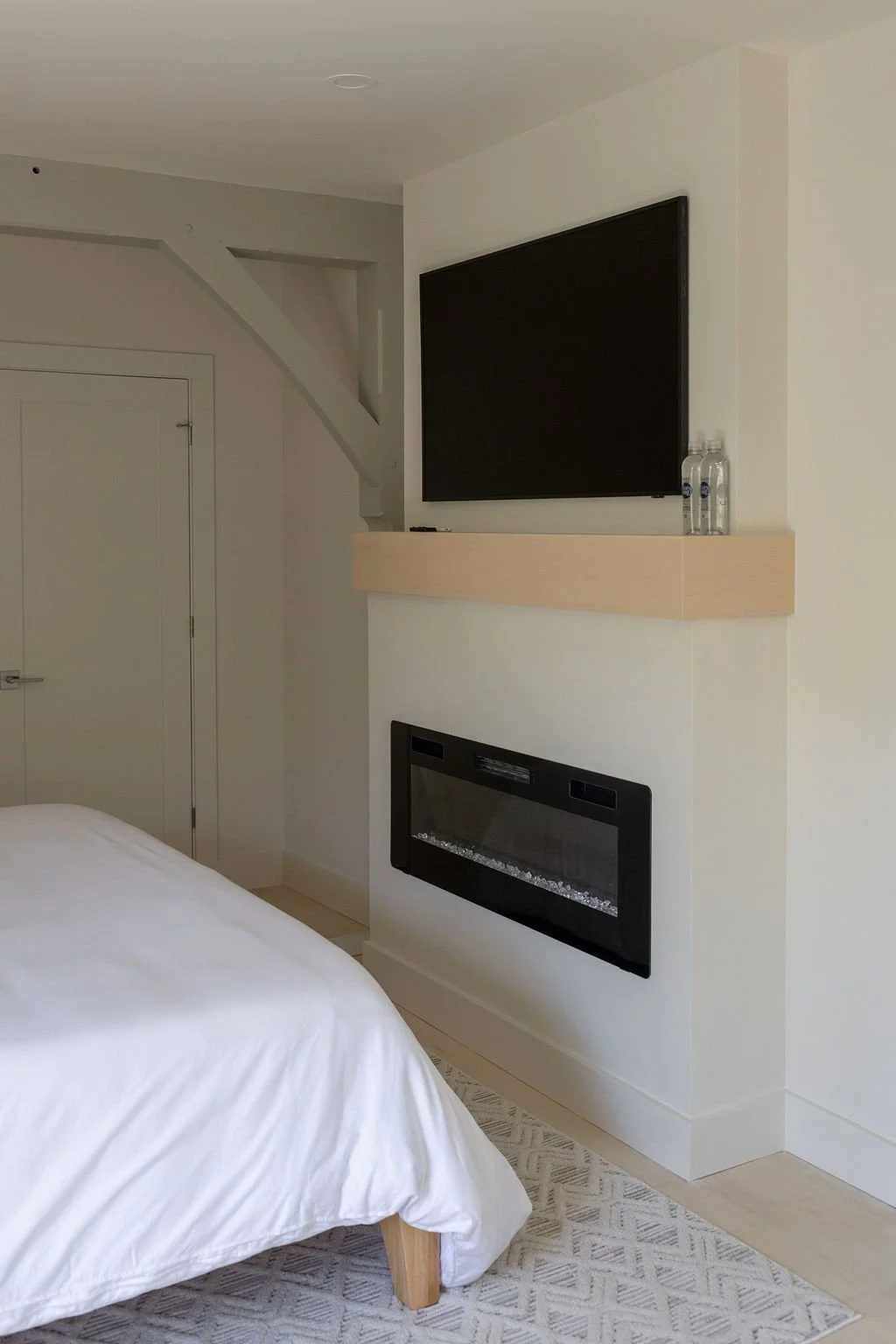 A corner of hotel bedroom with a bed covered in white bedding, a wall-mounted TV, a modern fireplace, and a light wood shelf with two water bottles on it.