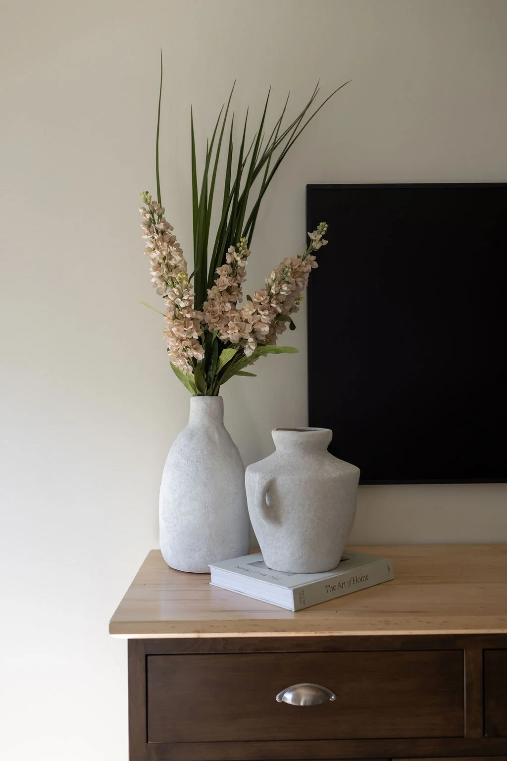 Decorative vases with pink flowers and green leaves on a wooden side table, next to a book titled "The Art of Home," with a black framed television on the wall behind.