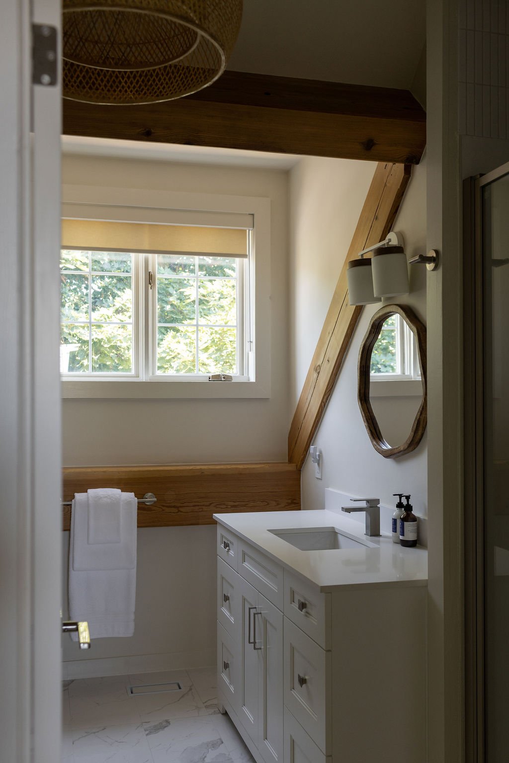 A small bathroom with a white vanity, a sink, and a mirror. A window with a view of trees, a wooden beam and trim, and a towel hanging on a bar.