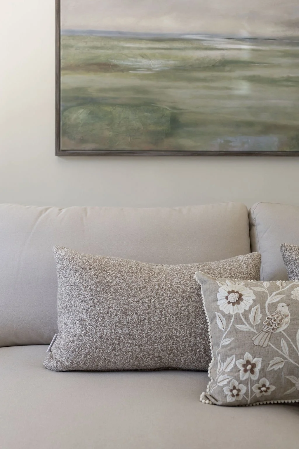 A beige couch with two decorative pillows, one textured in silver and the other with floral embroidery and a bird motif, in front of an abstract landscape painting on a white wall.