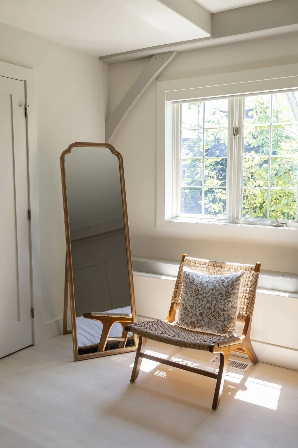 A sunlit corner of a hotel suite with a full-length mirror leaning against the wall, a wicker chair with a decorative pillow, a window allowing natural light, and a white door.