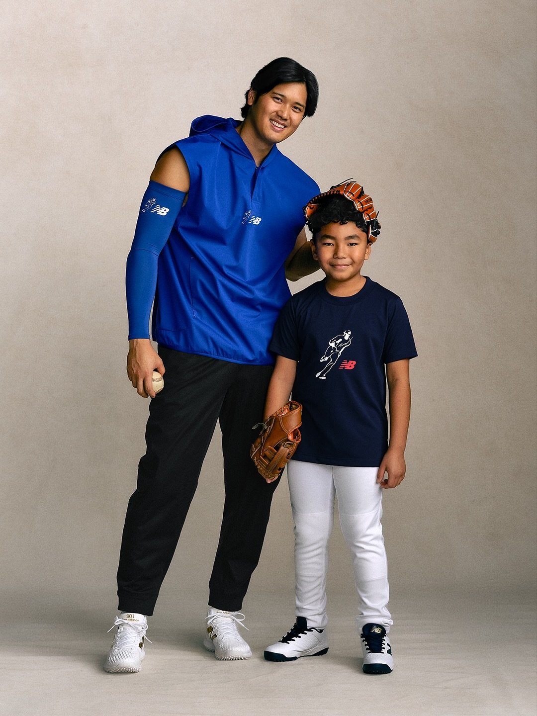 A man and a boy stand together in sports attire. The man wears a blue sleeveless top and black pants, holding a baseball and wearing a blue New Balance arm sleeve. The boy wears a dark blue t-shirt with a jumping figure logo, white pants, and holds a