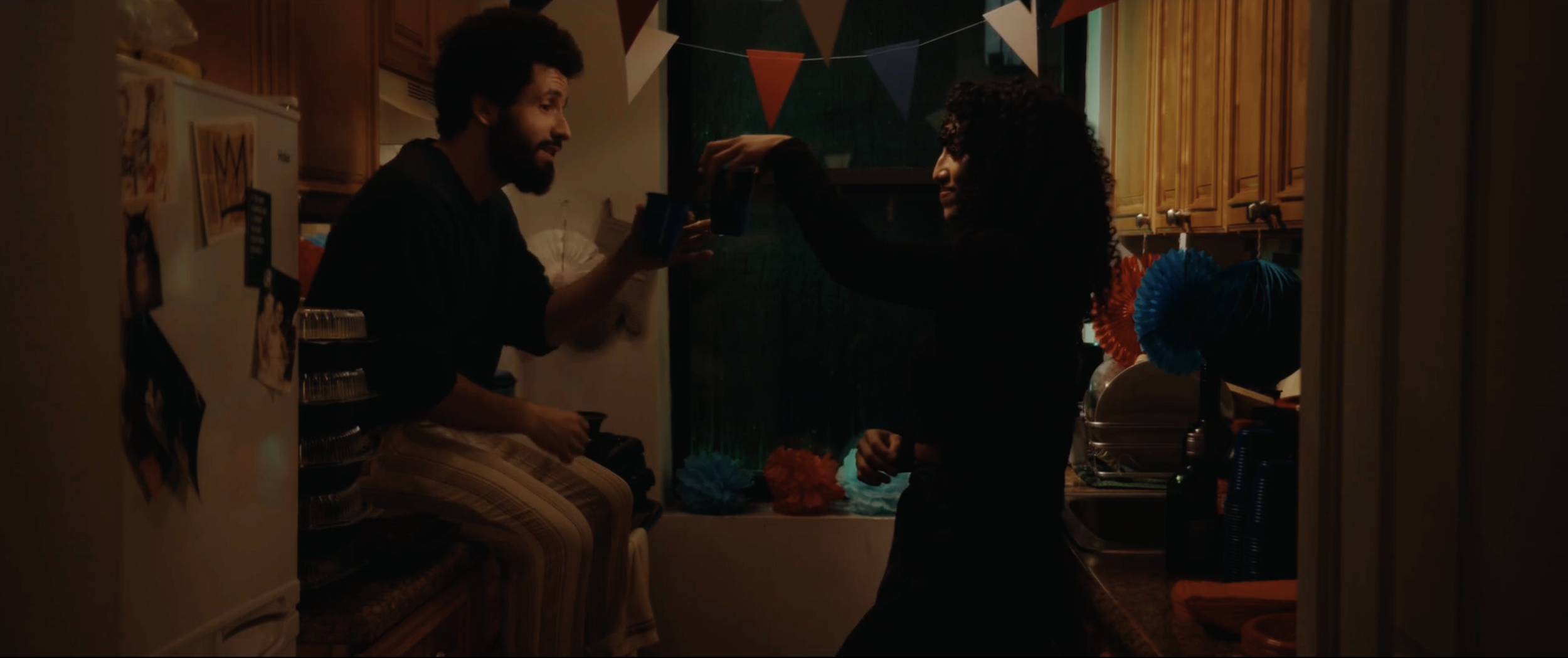 Two people celebrating with drinks in a dimly lit kitchen decorated with colorful bunting and paper pom-poms.