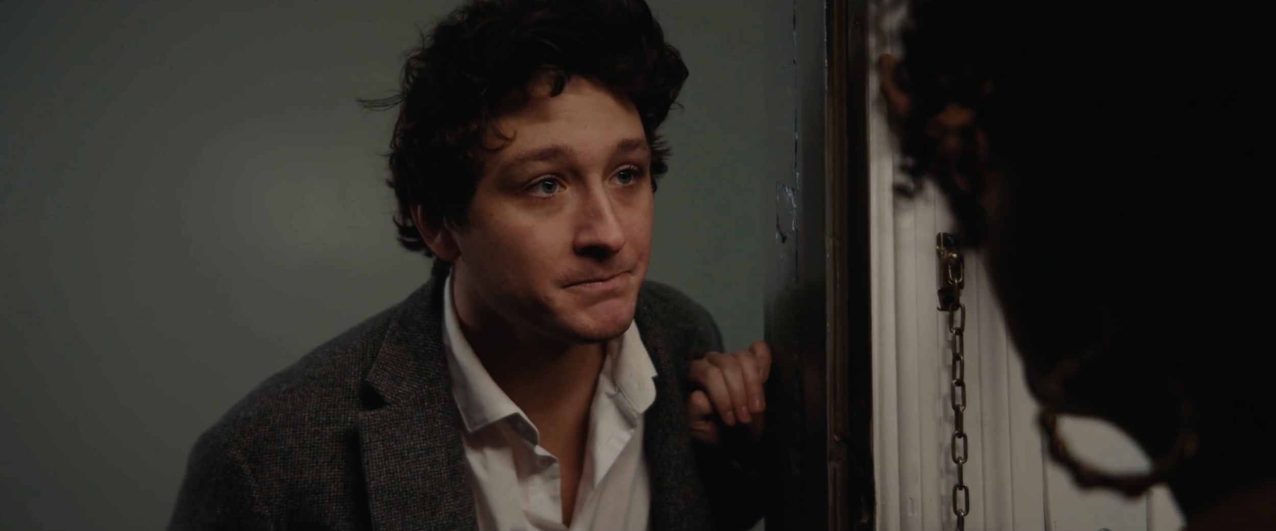 A man with curly hair and a serious expression, dressed in a white shirt and dark blazer, leaning close to another person at a door.
