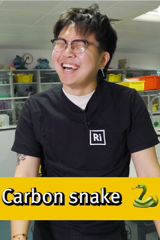 A person with short dark hair, glasses, and a black T-shirt with a periodic table element symbol, smiling or laughing in a laboratory setting.