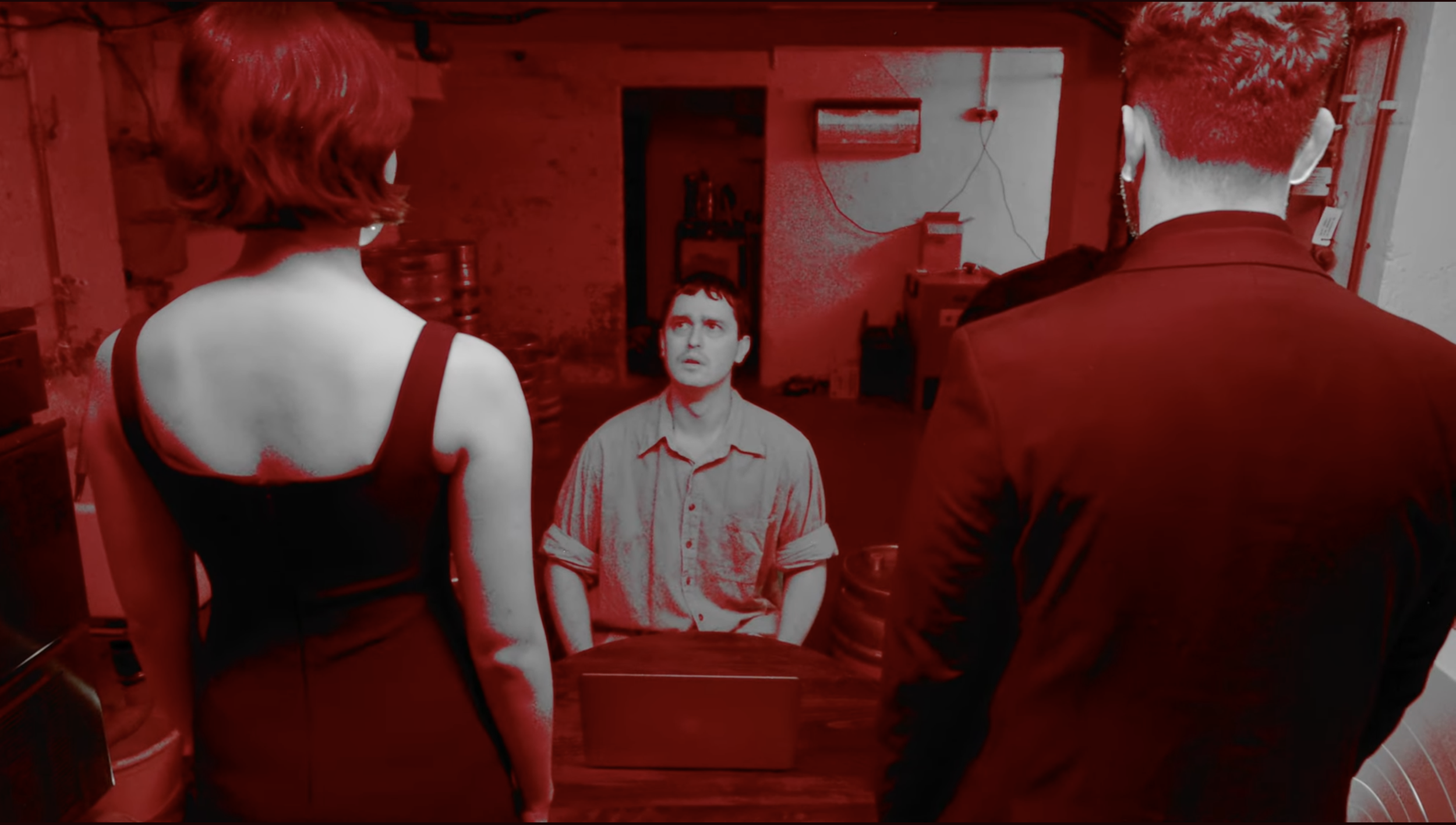 Three people are standing and one person is sitting in a room, having a conversation. The person sitting appears to be a young man with dark hair, wearing a light-colored shirt. The woman on the left has short, curly hair and is wearing a sleeveless top. The man on the right is seen from behind, has short hair, and is wearing a dark jacket. The room has visible background elements such as a wall with wires, a shelf, and some equipment or storage units.