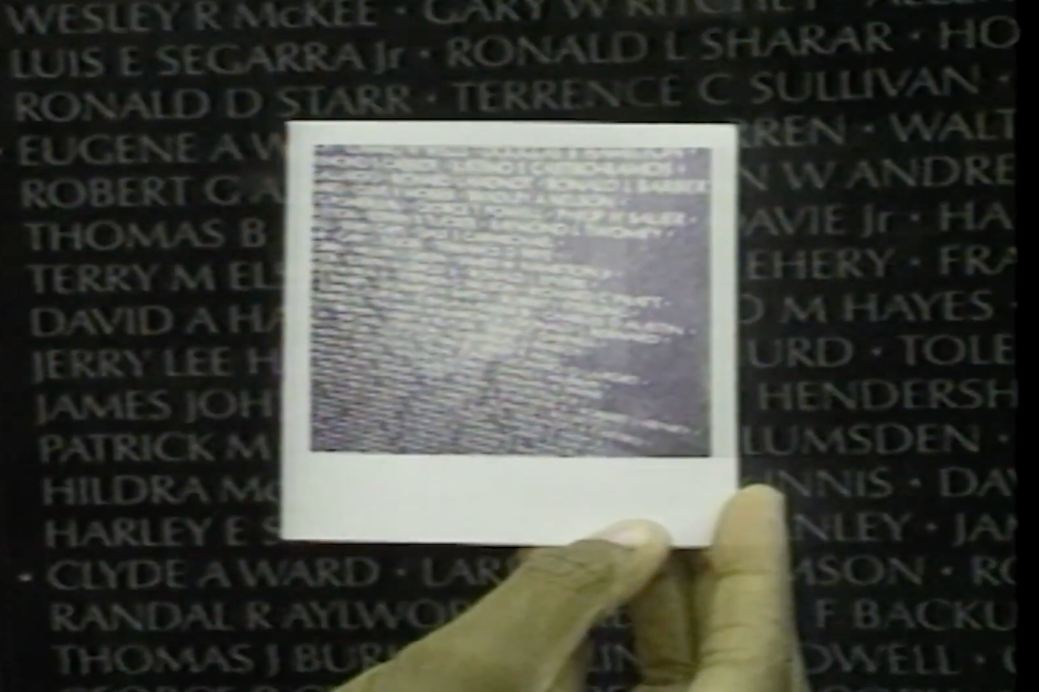 The image shows a person holding a framed photograph or document in front of a black wall with white names inscribed on it. The photograph or document appears to contain text that is difficult to read due to blurriness.