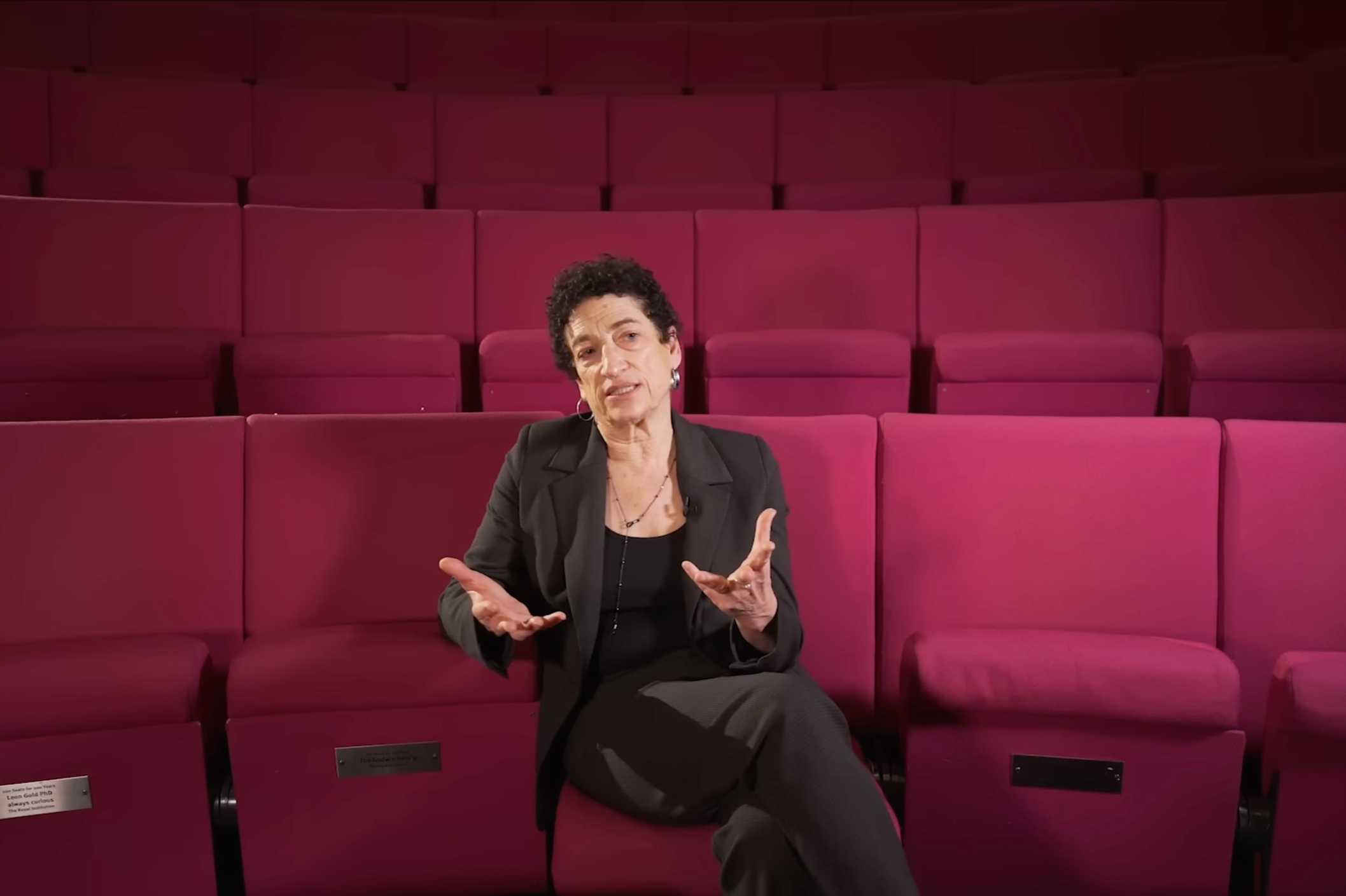 Woman with short curly hair and earrings seated in an auditorium with red seats, speaking and gesturing with her hands.