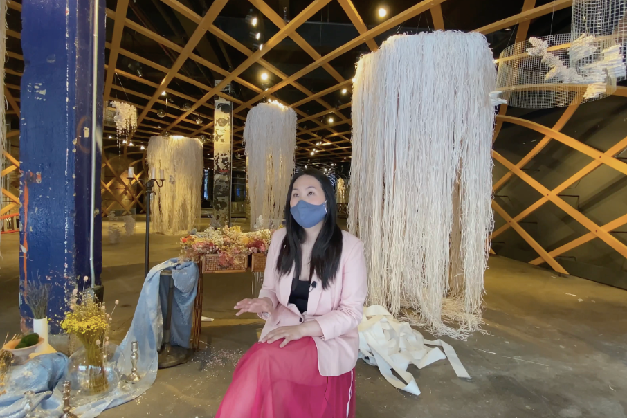 A woman wearing a face mask sitting in front of large hanging textile art installations inside a decorated space.