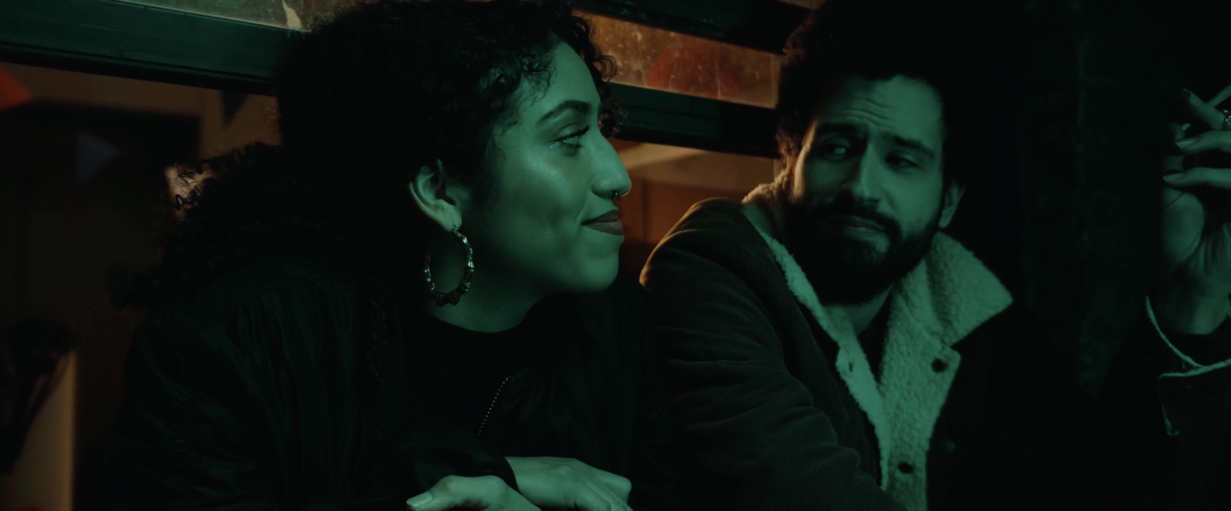 Two people sitting close together inside a dimly lit establishment, engaging in conversation. The woman is on the left, with curly hair and hoop earrings, looking at the man. The man is on the right, with dark curly hair and a beard, holding a cigarette.